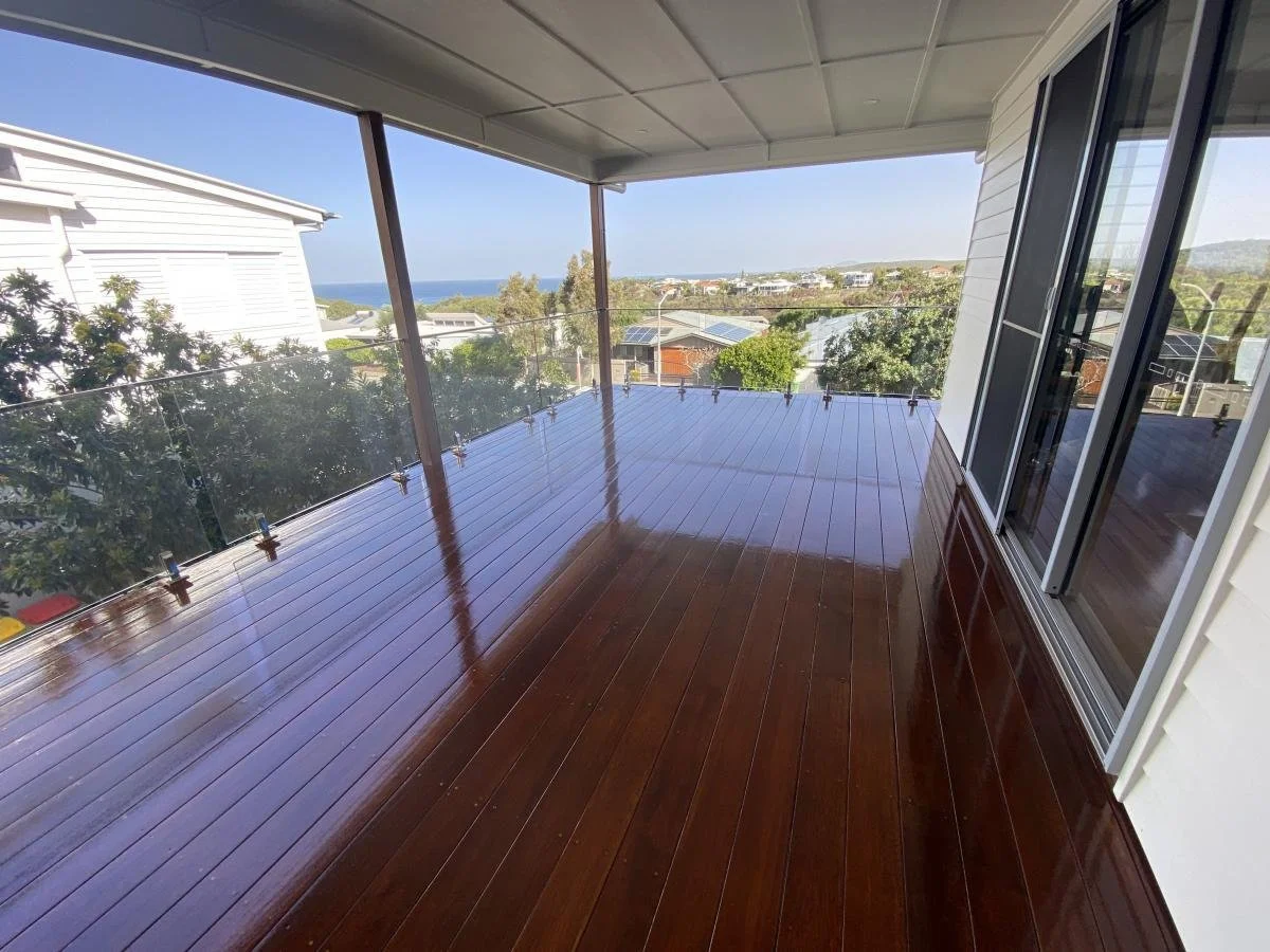 Empty balcony with polished wooden flooring, glass railing, sliding glass door, and view of trees, rooftops, and the ocean in the distance.