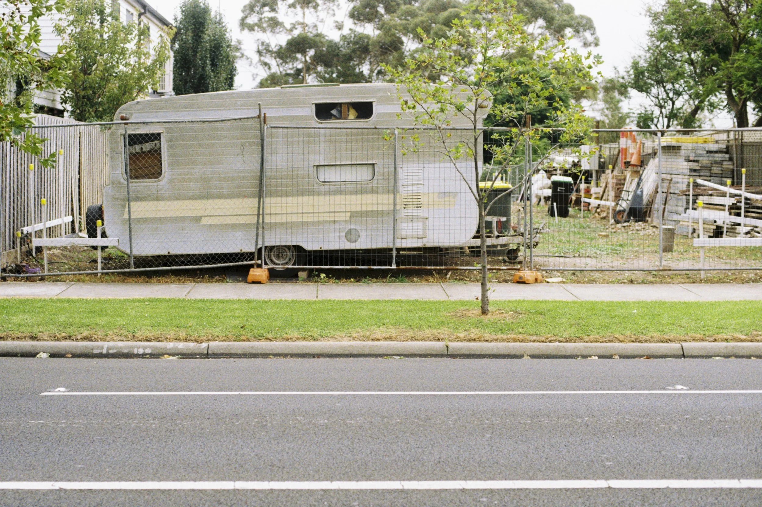 A residential street with a curb, a grassy lawn, and a tree in the foreground. Behind the fence, there is a vintage camper trailer surrounded by a construction site with various materials, tools, and orange traffic cones. In the background, there are