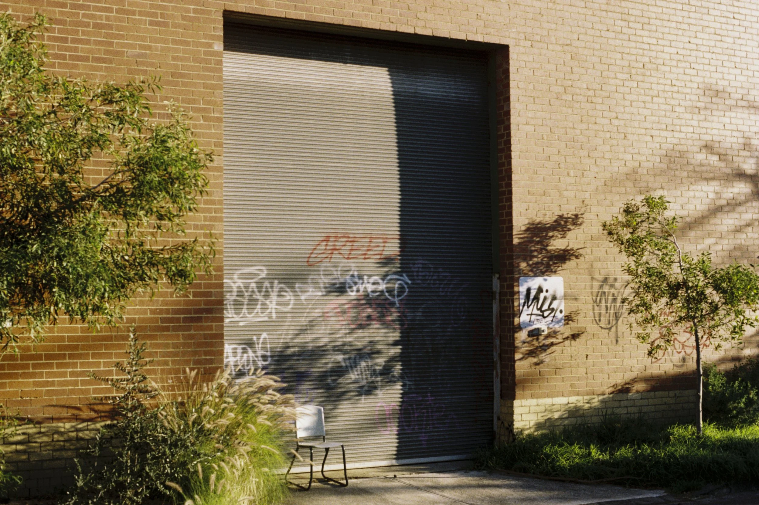 A brick building with a grey metal roll-up door covered in graffiti, flanked by small trees and bushes, with a white chair on the sidewalk in front.