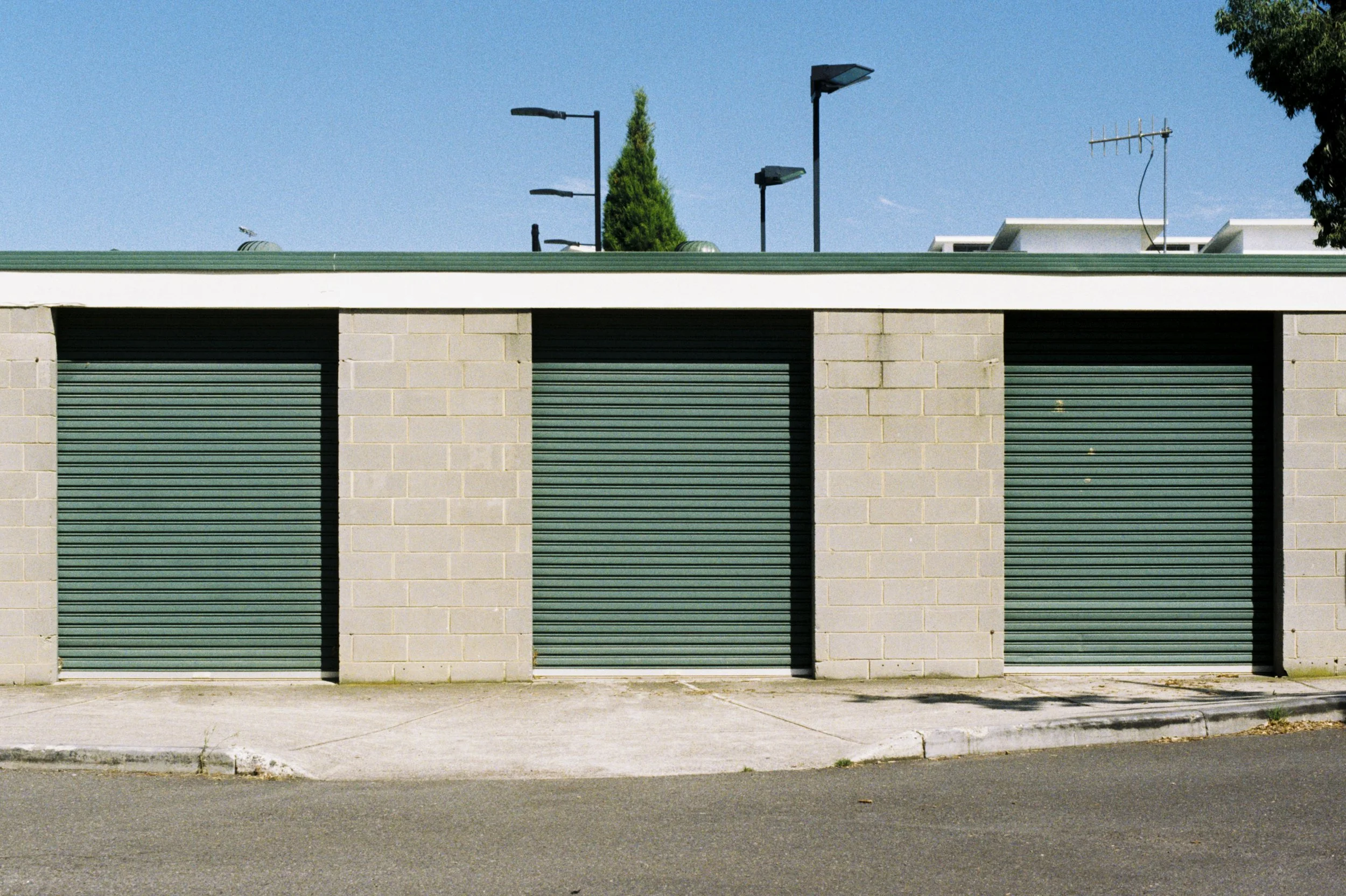 A building with three green garage doors, concrete block walls, a green roof, and a sidewalk in front. The sky is clear and blue, with some trees and rooftop equipment visible.