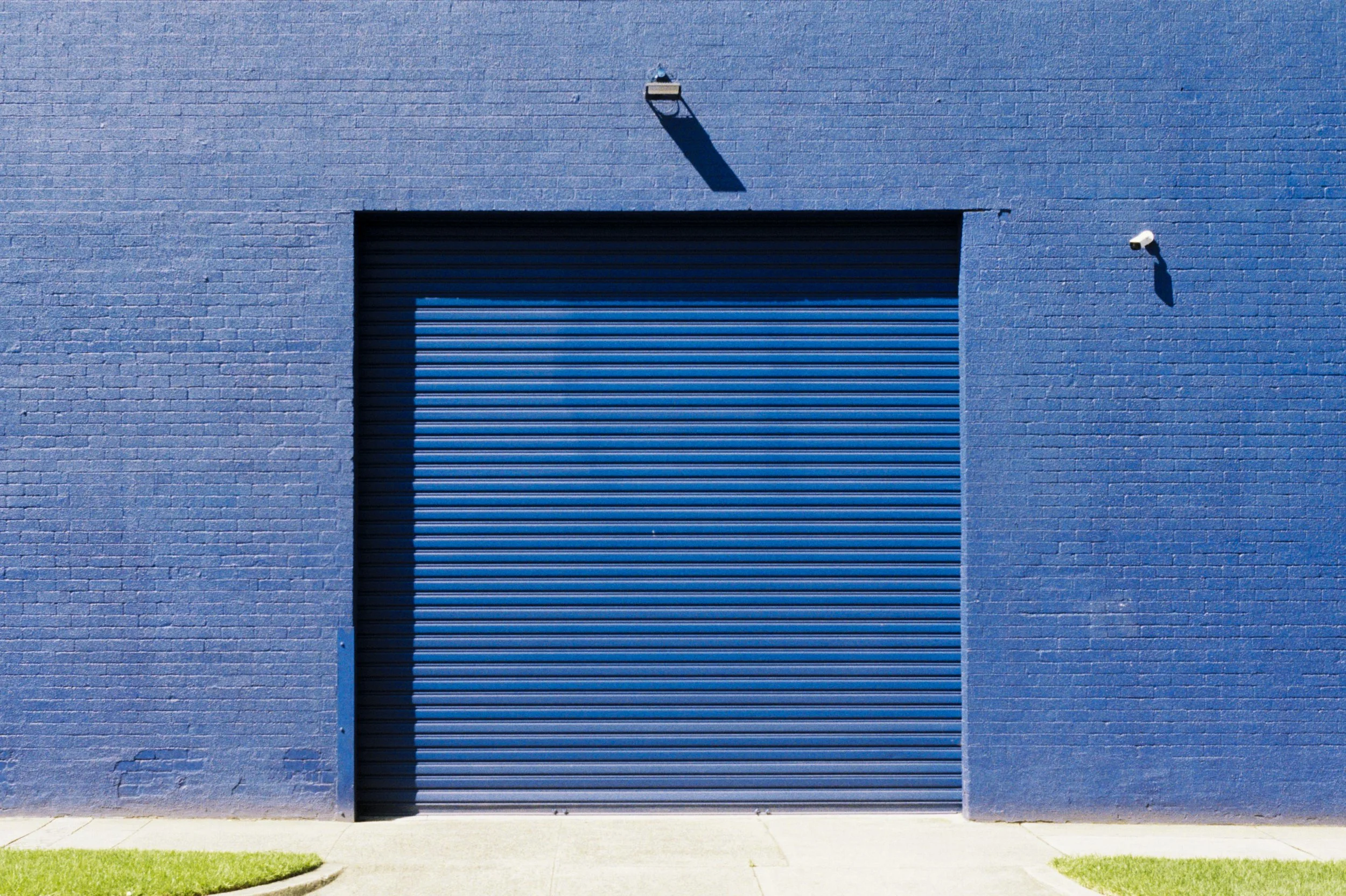 Blue building with a closed metal roll-up door, two security cameras, and a light fixture, with a concrete sidewalk and small patches of grass in front.