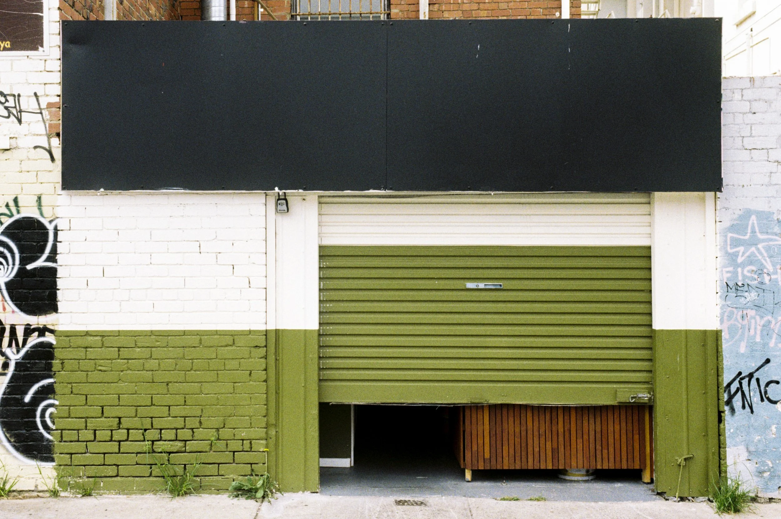 A building with a large black signboard above a green metal shutter door. Part of a brick wall with graffiti on the left and right sides.