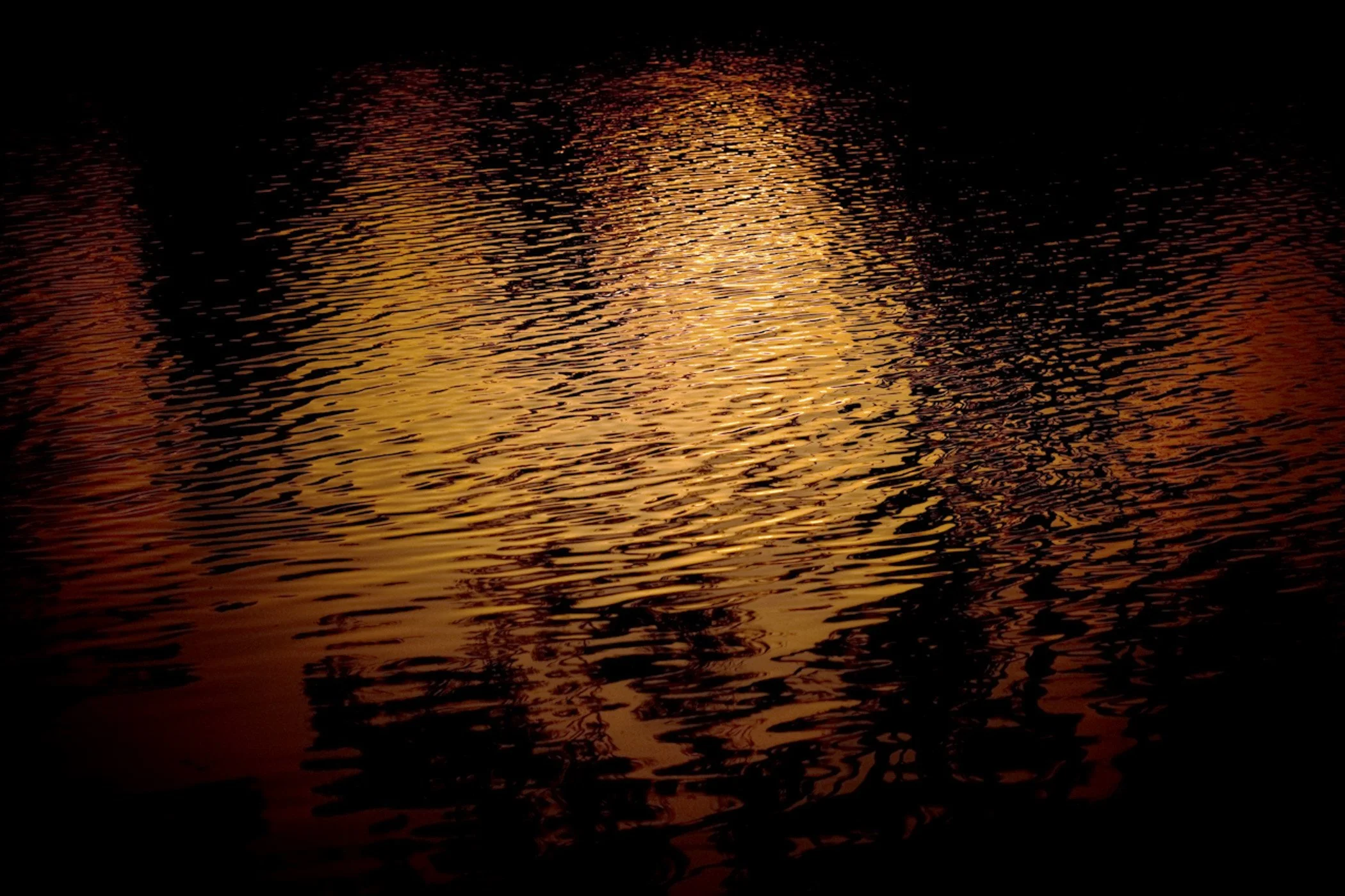 Reflection of a sunset on a calm body of water, creating warm orange and gold hues with ripples.
