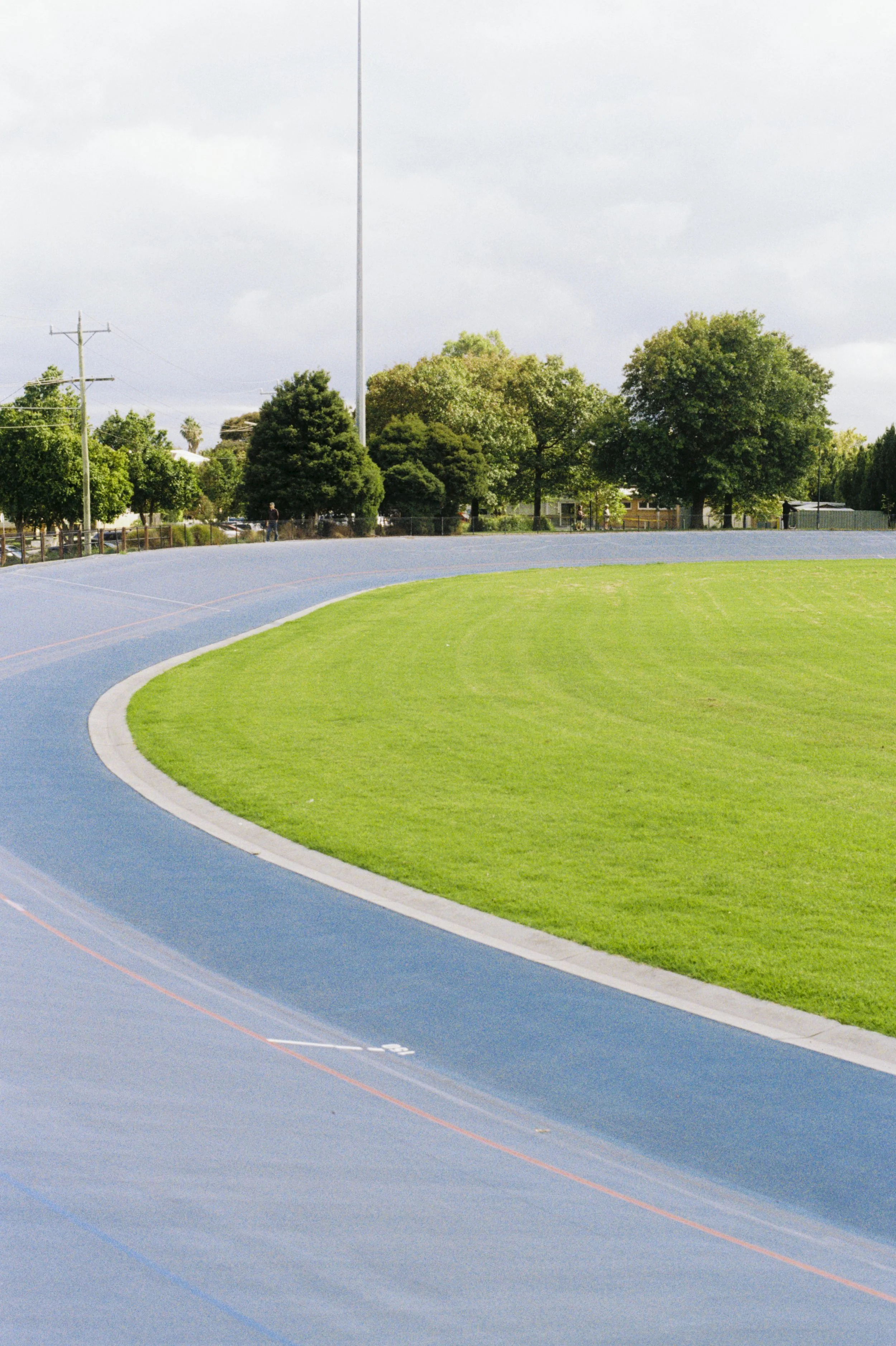 An outdoor running track with a blue surface, green grass in the infield, and trees in the background under a cloudy sky.