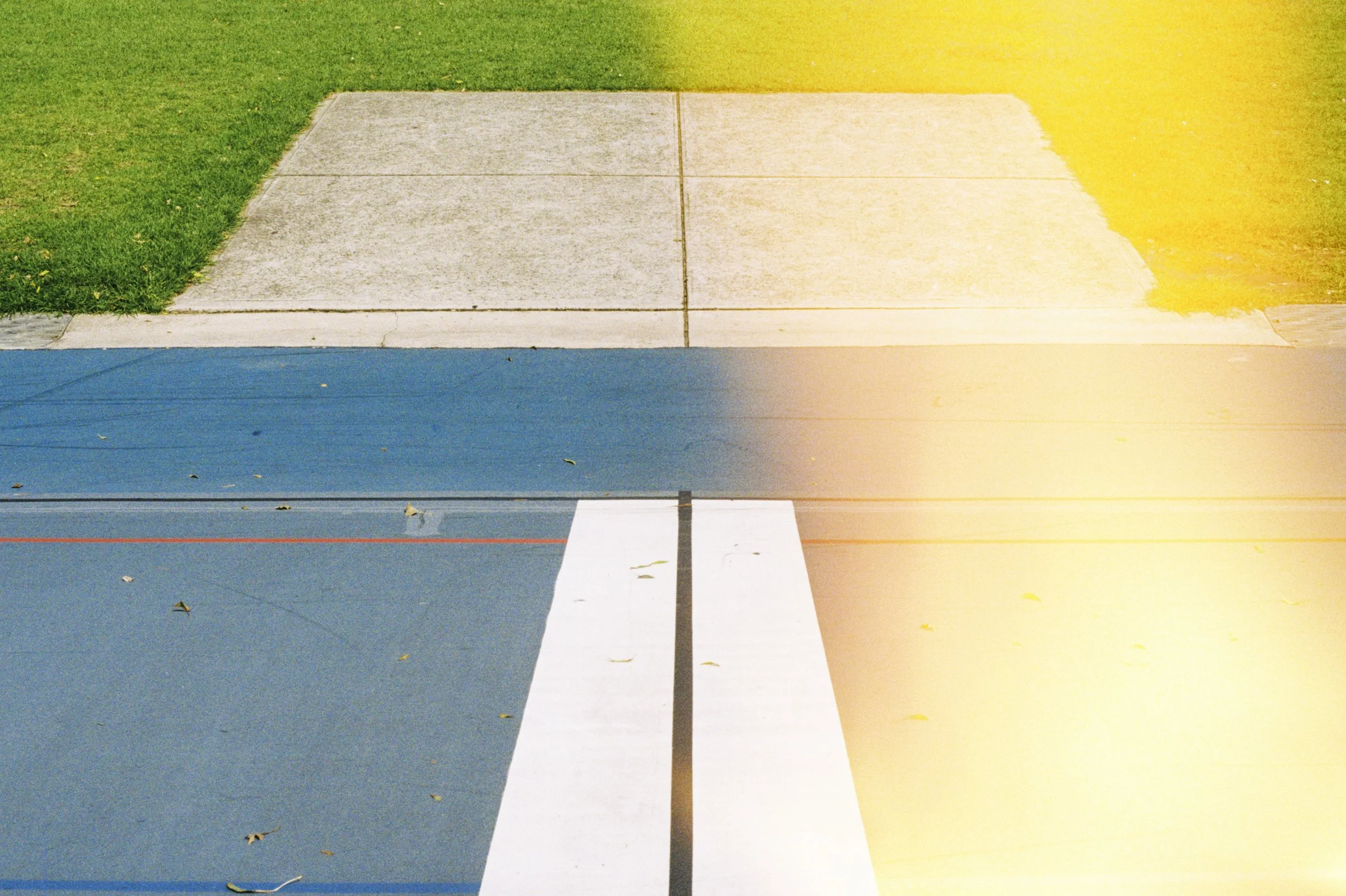 An outdoor scene showing a sidewalk with three sections, a strip of green grass to the left, and a yellowish area to the right. The sidewalk is divided into four concrete slabs, with some leaves scattered on the blue section at the bottom and small c