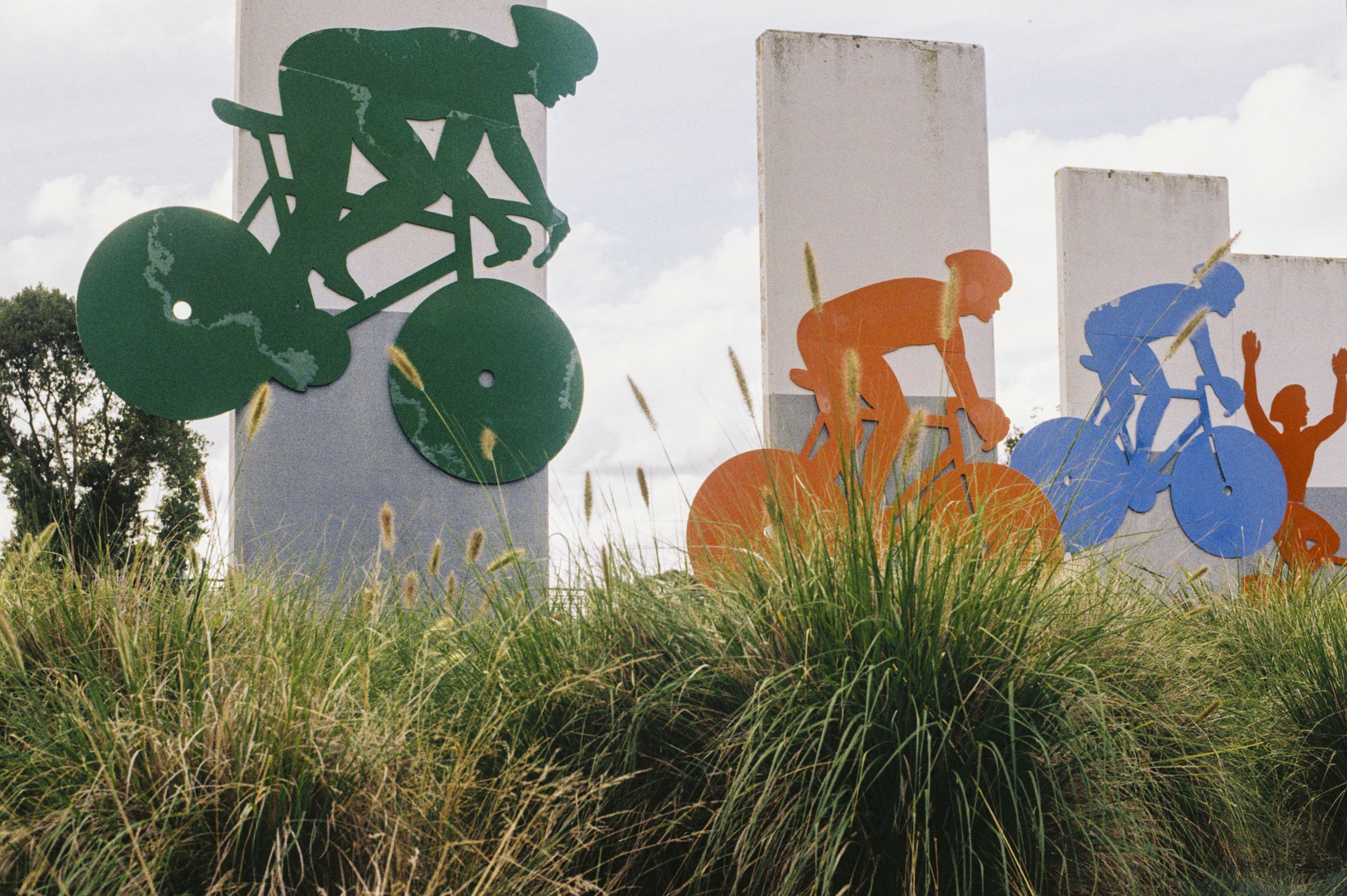Colorful sculptures of cyclists and a person raising their arms, set against tall white pillars, with grass in the foreground and a cloudy sky in the background.