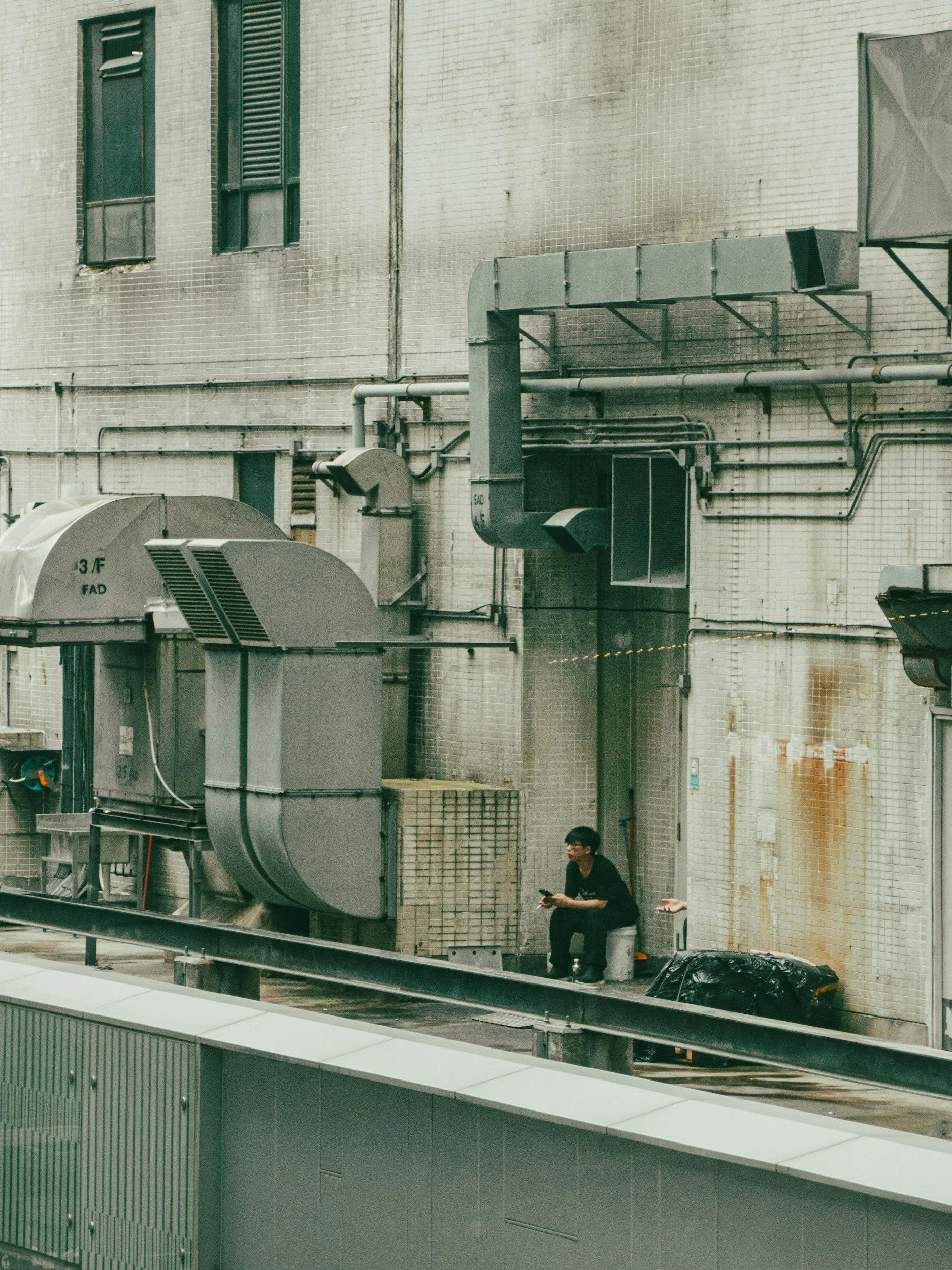 A person sitting on a small stool with a laptop, on a rooftop or industrial area, surrounded by large industrial ventilation and piping systems against a beige brick building wall.