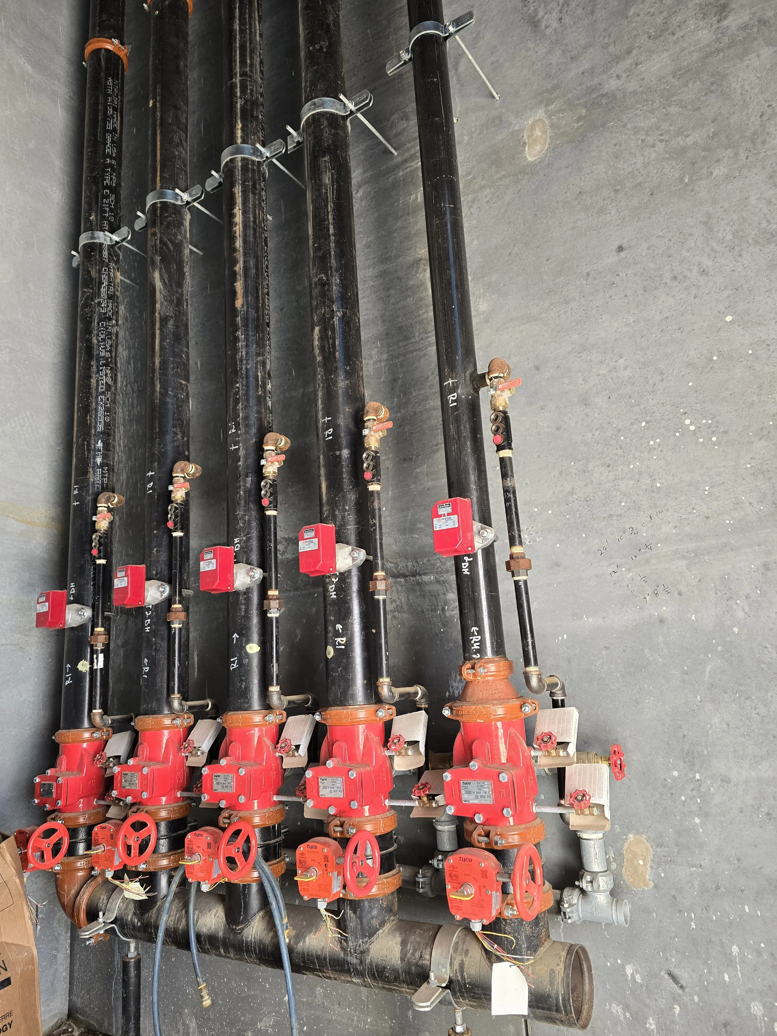 Five large black water pipes with red valves and digital meters, connected to smaller black pipes with red handwheels, on a concrete floor.