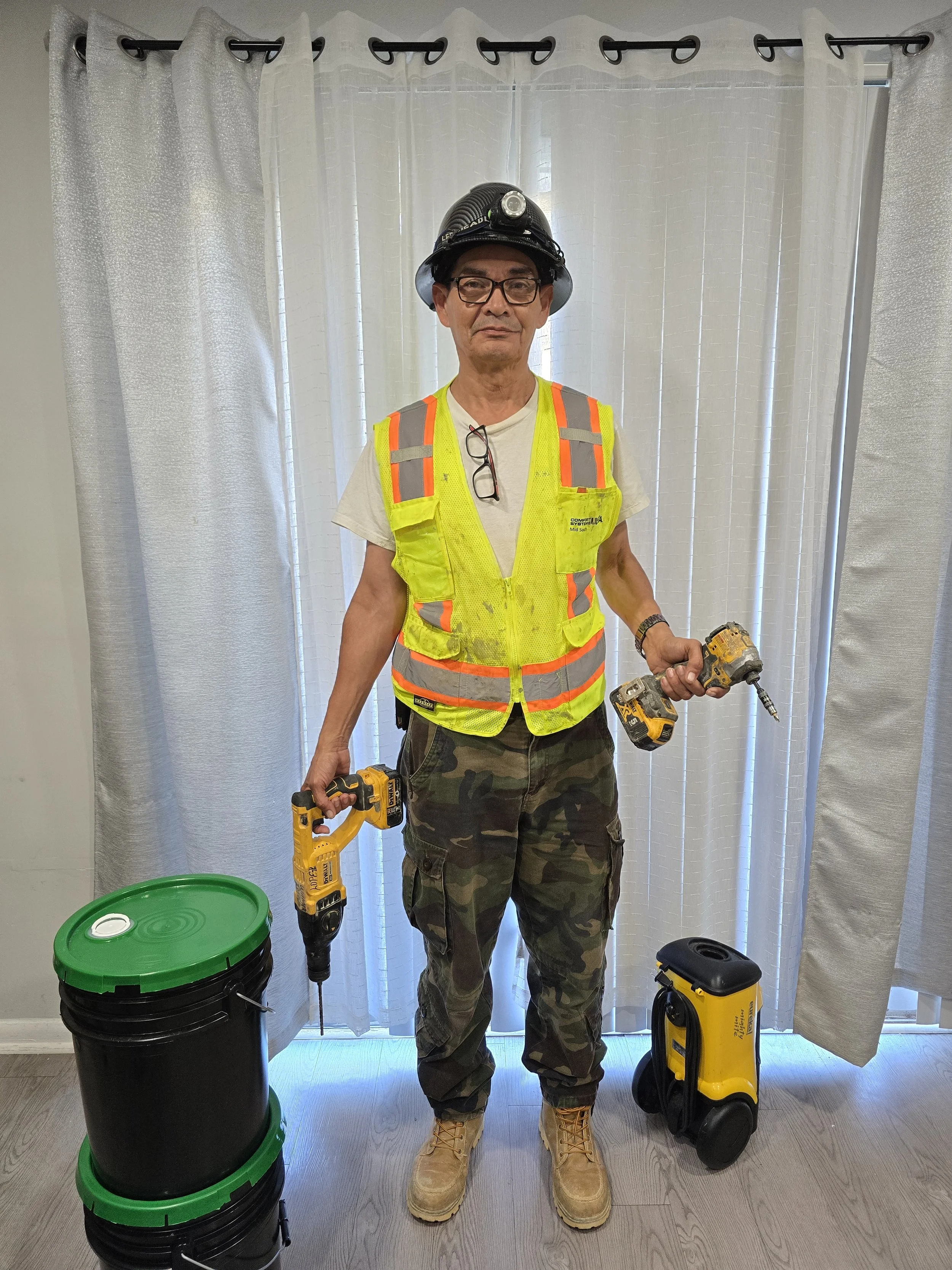 A man dressed as a construction worker wearing a yellow safety vest, camouflage pants, tan boots, and a black hard hat with a headlamp. He is holding a cordless drill in each hand and stands in front of white curtains. There are two yellow portable foggers on the floor beside him and two black buckets with green lids.