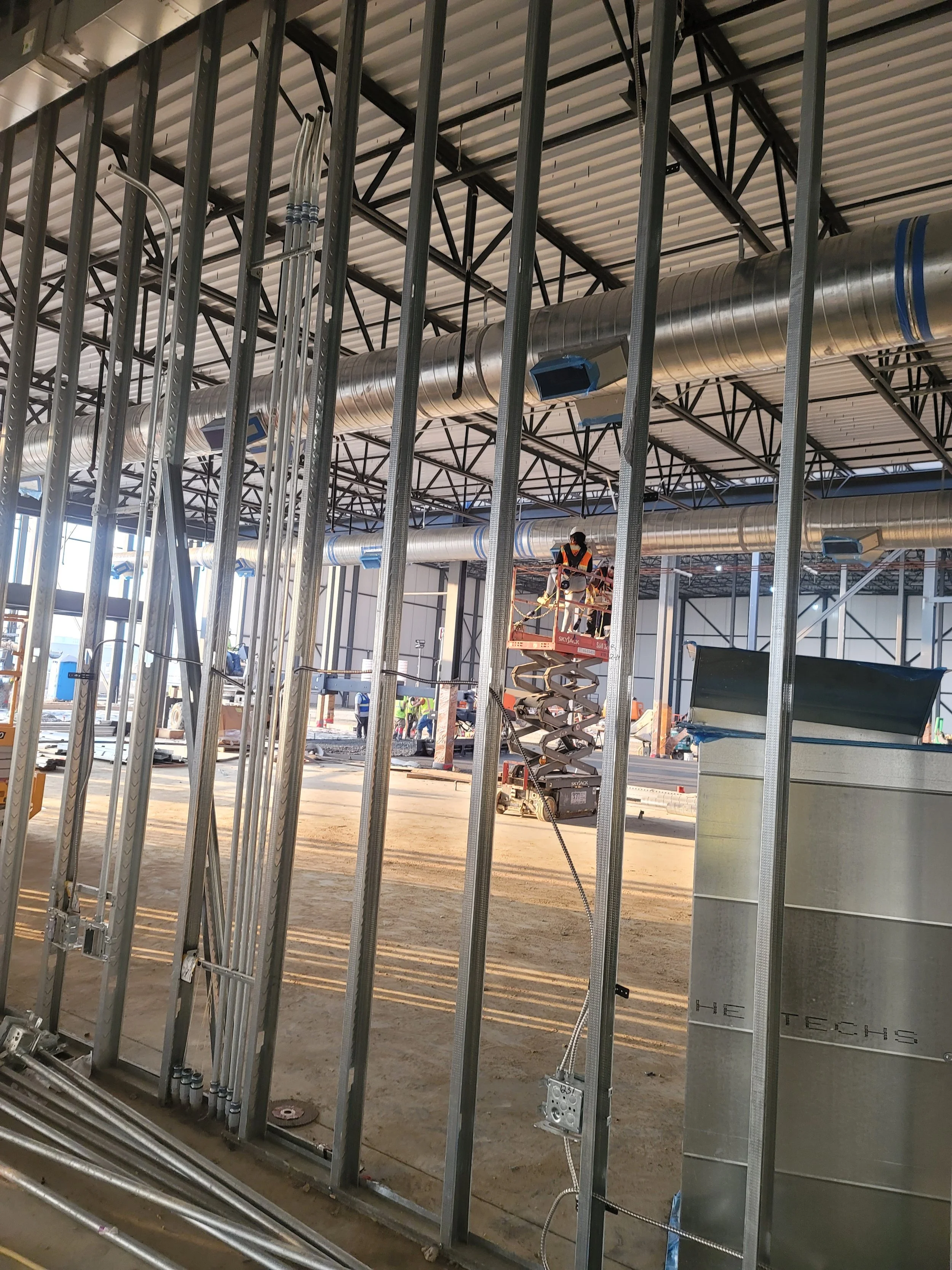 Construction site with metal framing, large ventilation ducts, and workers on a lift installing equipment inside a building.