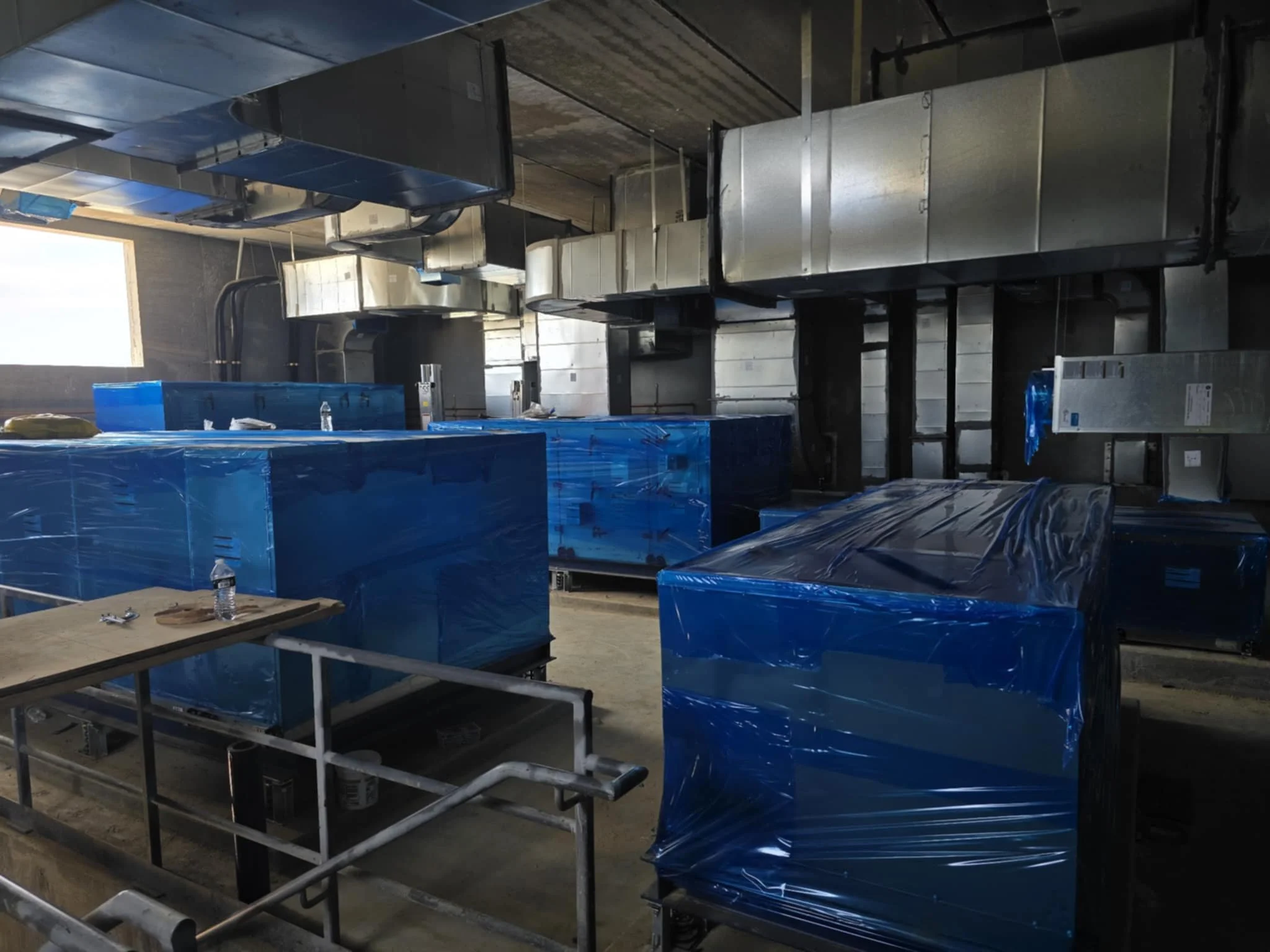 Industrial kitchen or commercial food preparation area with large equipment covered in blue plastic wrap, metal ventilation ducts, stainless steel counters, and a window allowing natural light.