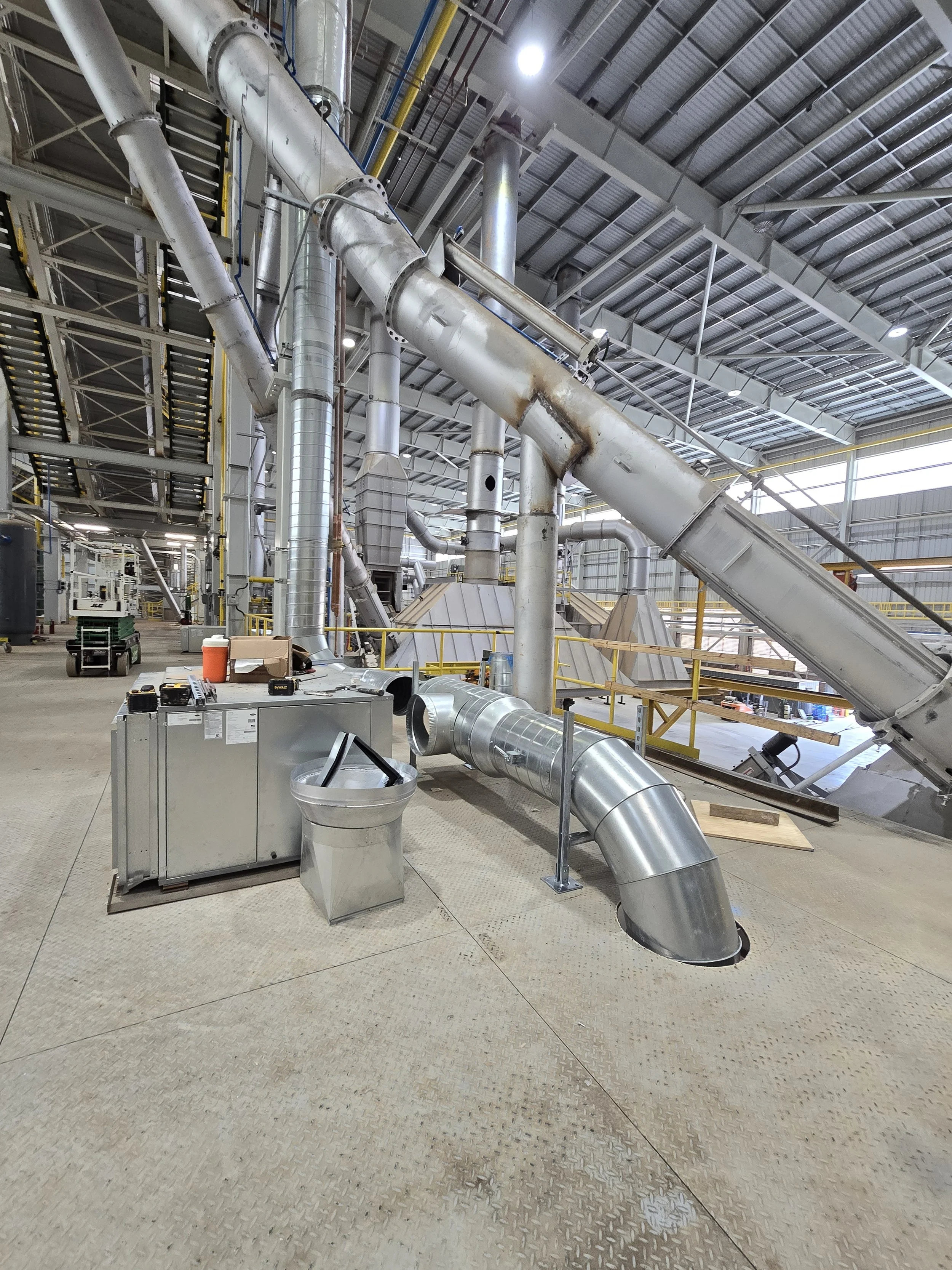 Industrial manufacturing plant with large metal pipes and ducts, some rusted, running overhead and across the floor. There is a small metal table with various items on it, including a plastic bucket, and the floor is textured metal with some dirt sta