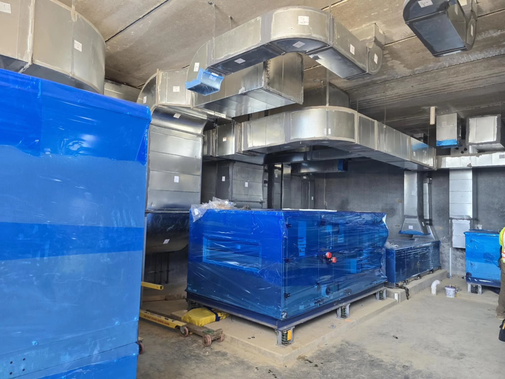 Construction site with exposed HVAC ductwork and machinery, some wrapped in blue protective plastic, on a concrete floor with a worker in safety gear visible on the right side.