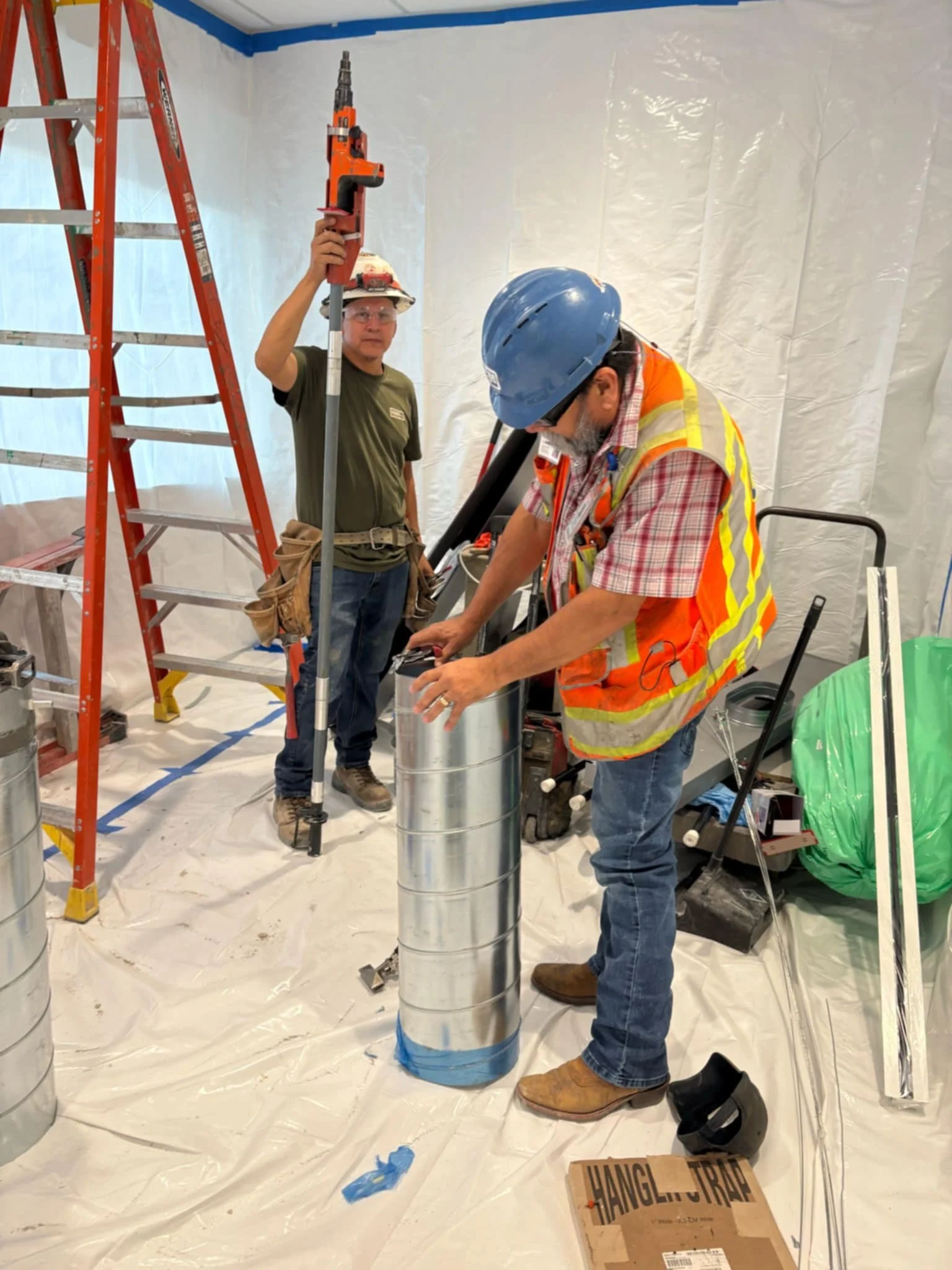 Two construction workers in safety gear work inside a white plastic-walled room. One worker wearing an orange safety vest and blue hard hat is handling a cylindrical metal duct. The other worker, wearing a helmet and t-shirt, is standing next to a red ladder, holding a long pole with a tool attached.