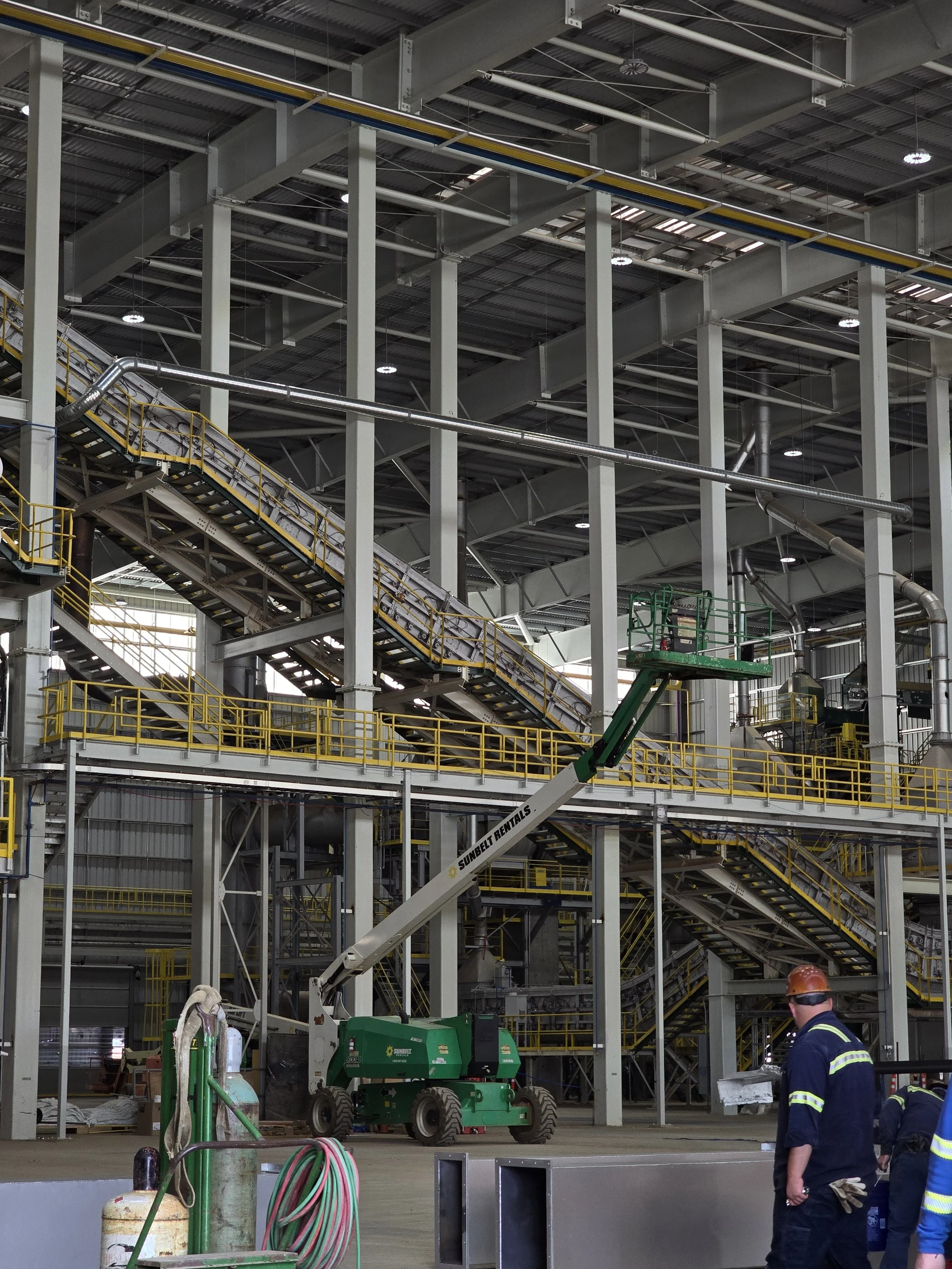Inside an industrial warehouse with steel structures, stairs, and walkways. A green lift is positioned near the center, with mechanical equipment and workers wearing hard hats and safety vests in the foreground.