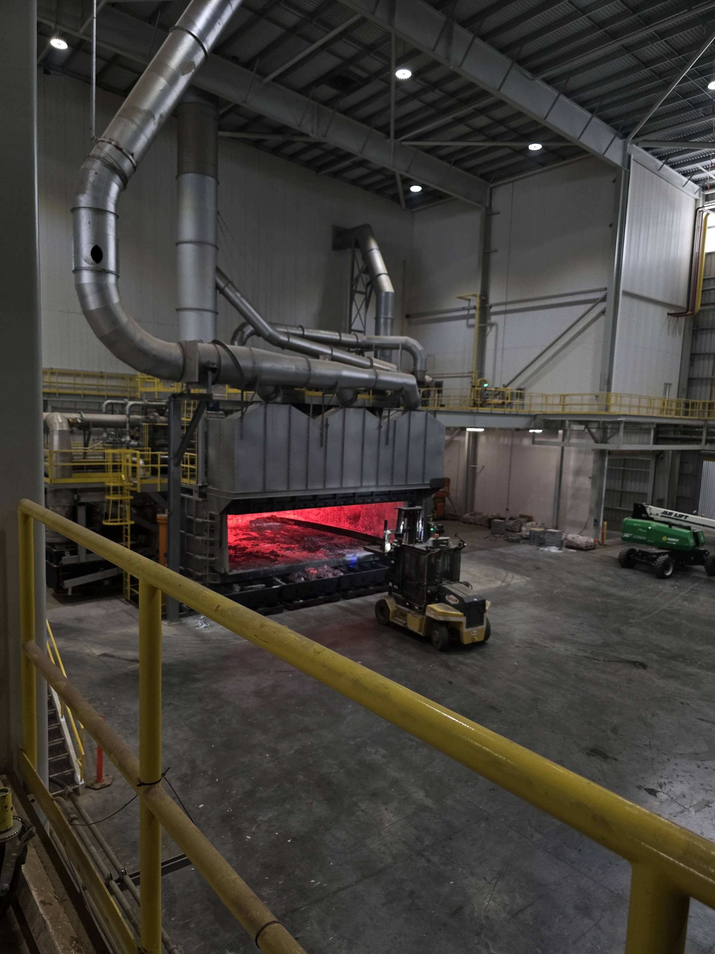 Industrial furnace in a factory with a yellow safety railing in the foreground and a forklift operating near the glowing red furnace.