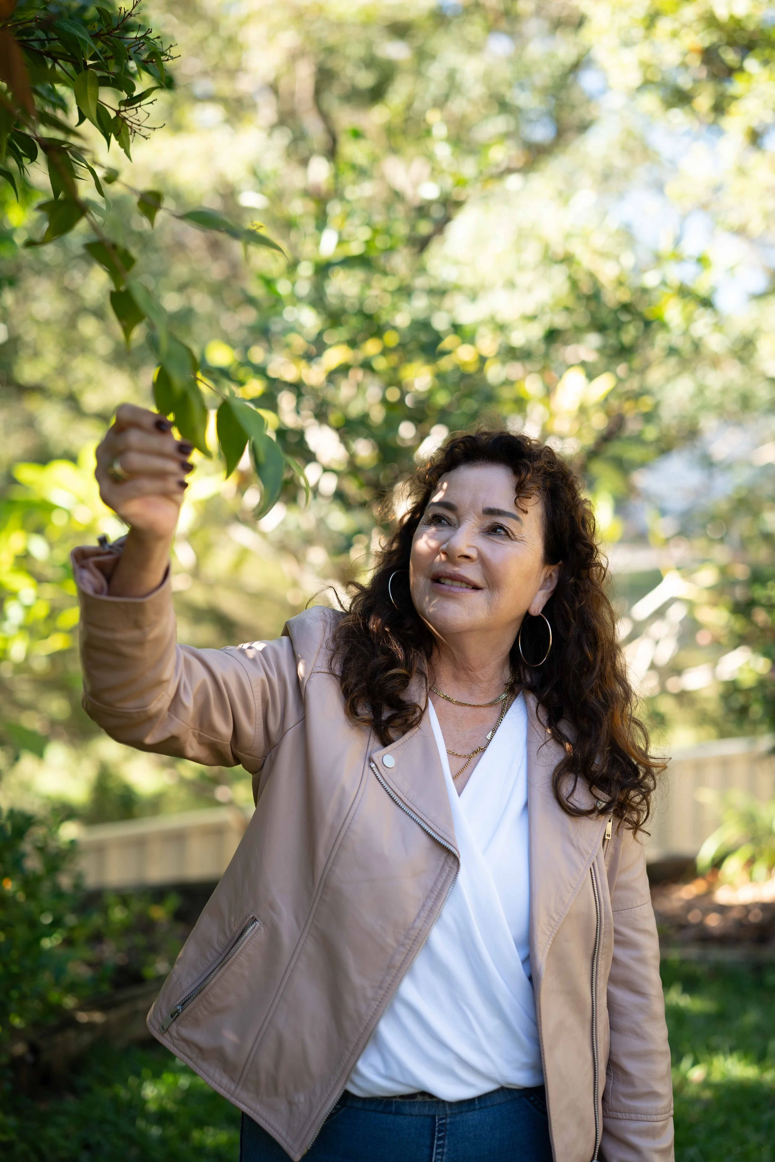 Sue Dalby, Senior Counselling Psychologist, wearing a beige jacket and white shirt, stands outdoors in a garden, holding a plant branch with green leaves.