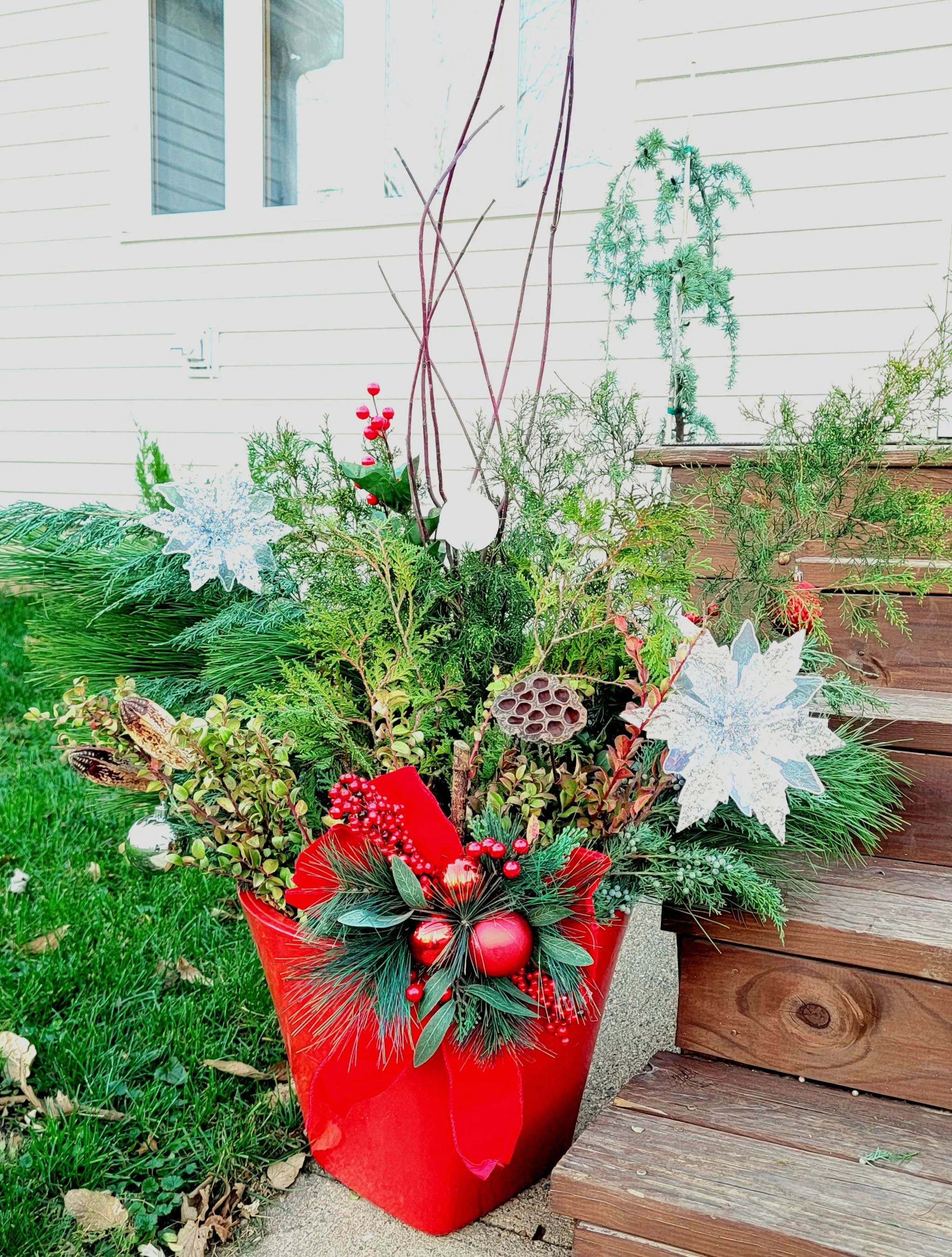 Festive outdoor Christmas arrangement with ornaments, pine branches, and decorative elements in a red container, placed next to wooden stairs on a porch.
