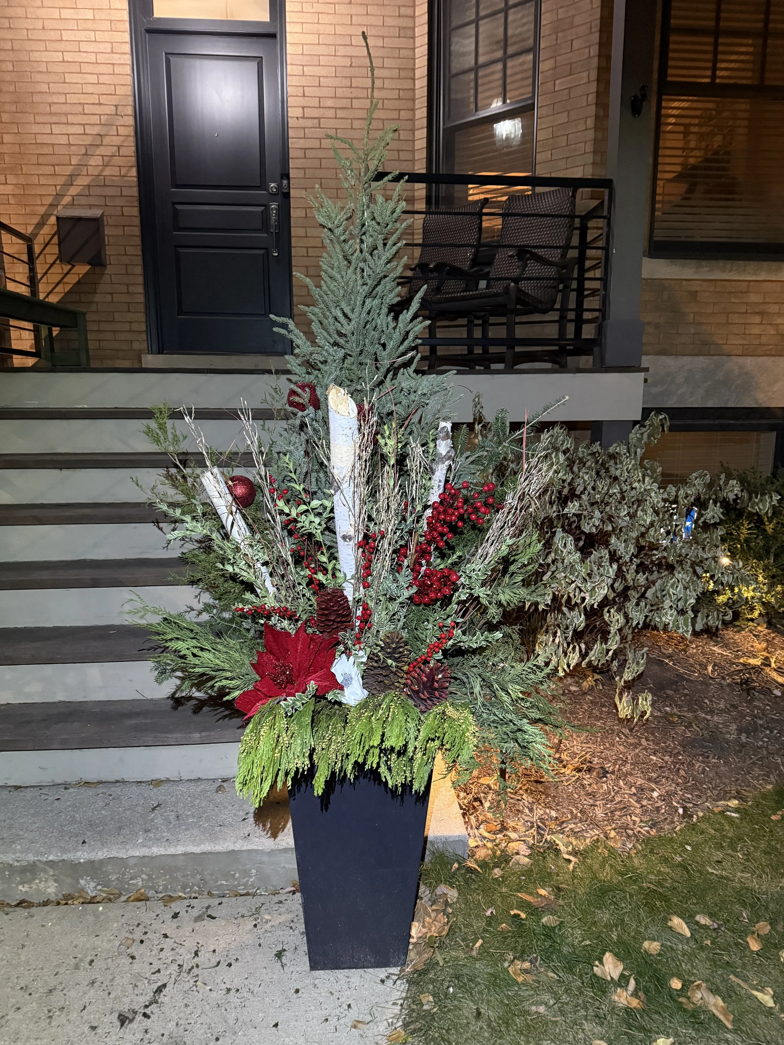 Decorative Christmas floral arrangement with greenery, red poinsettias, pine cones, red berries, and ornaments in a black planter outside a brick house.