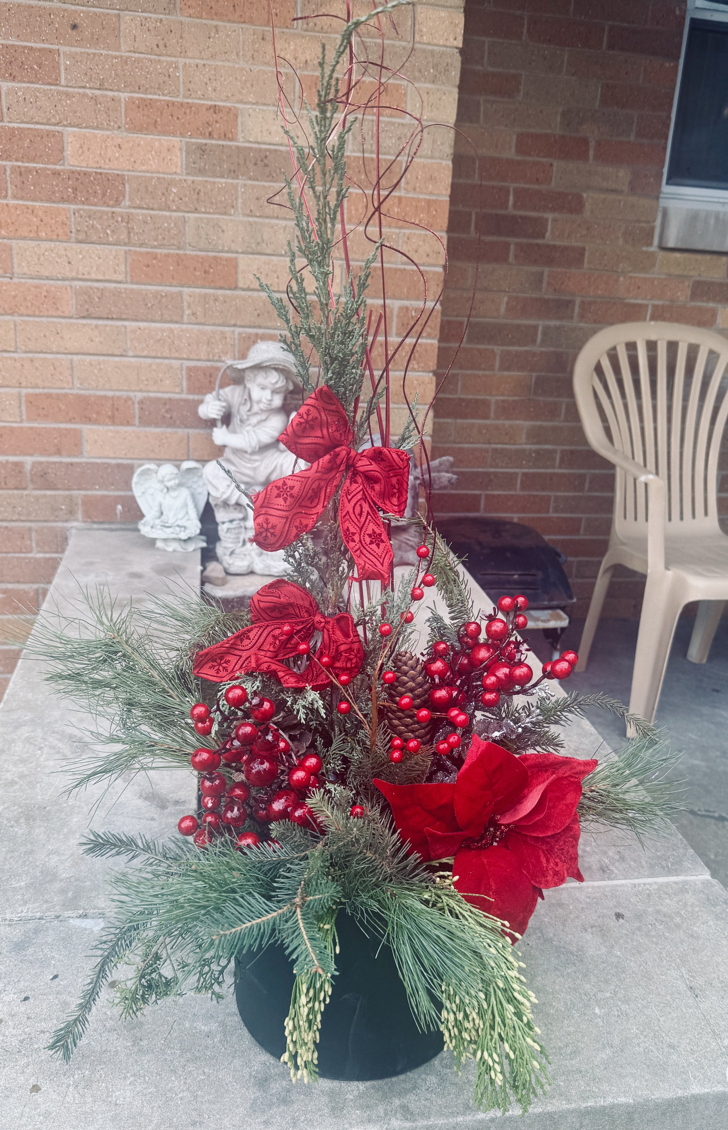 A holiday floral arrangement with red ribbons, berries, pinecones, and evergreen branches on a table outside, with Christmas-themed decorations and a brick wall in the background.