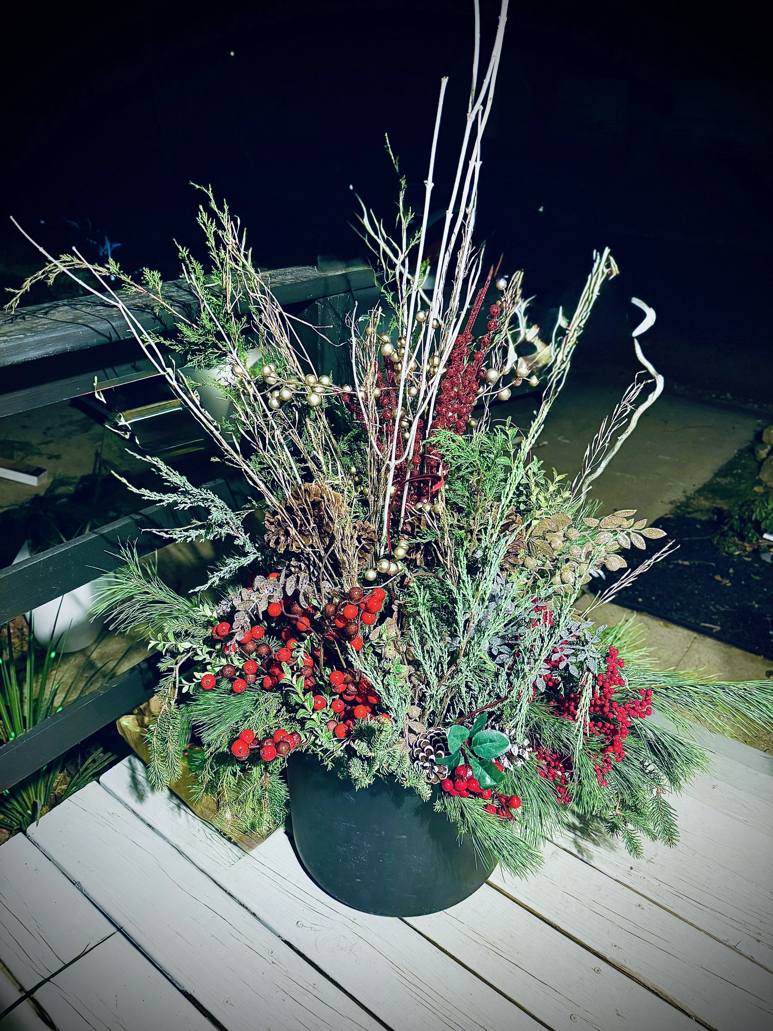 Holiday floral arrangement with evergreen branches, red berries, pinecones, dried branches, and gold ornaments in a black pot on a white wooden table at night.