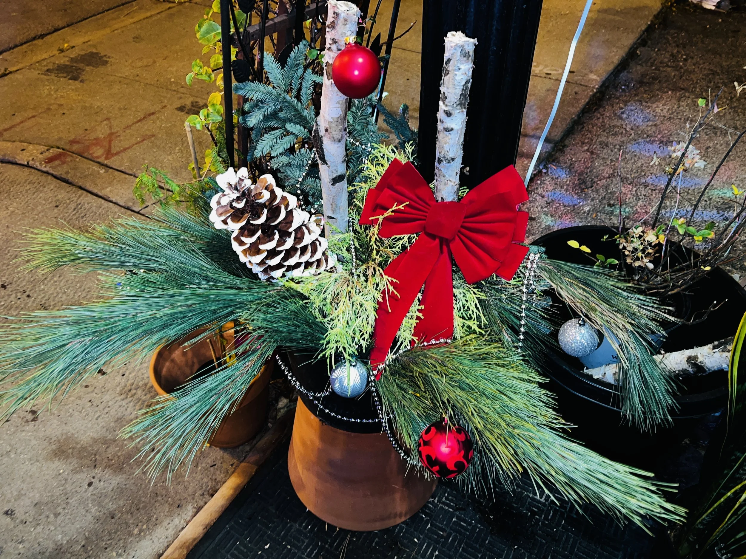 Decorative Christmas arrangement in a planter with pine branches, a pine cone, a red bow, silver and red ornaments, and small trees inside planters near a sidewalk.