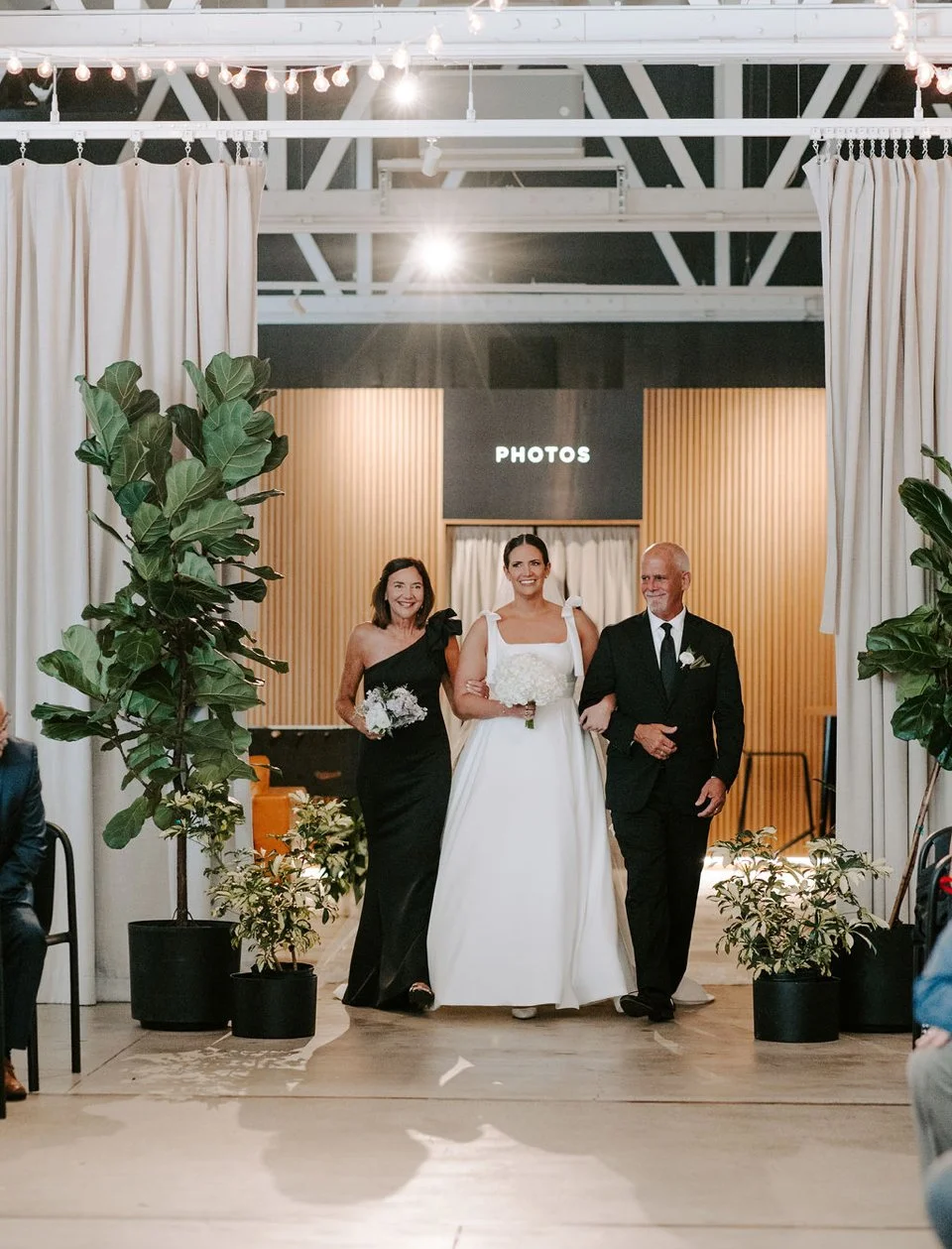 A bride walking down the aisle with her parents during her wedding ceremony, surrounded by potted plants and decorated with curtains and string lights.