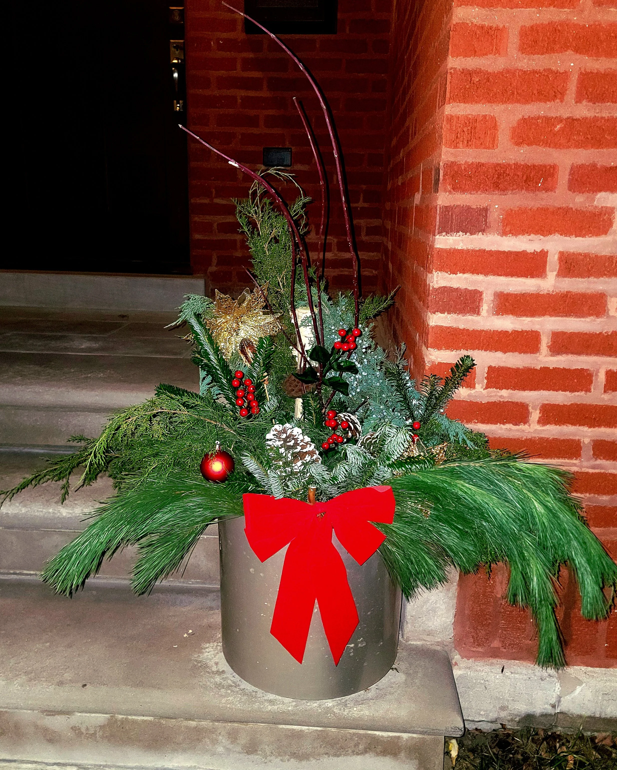 Christmas floral arrangement in a gray pot with a red bow, pine branches, red ornaments, berries, and decorative sticks, placed on steps outside near a brick wall.