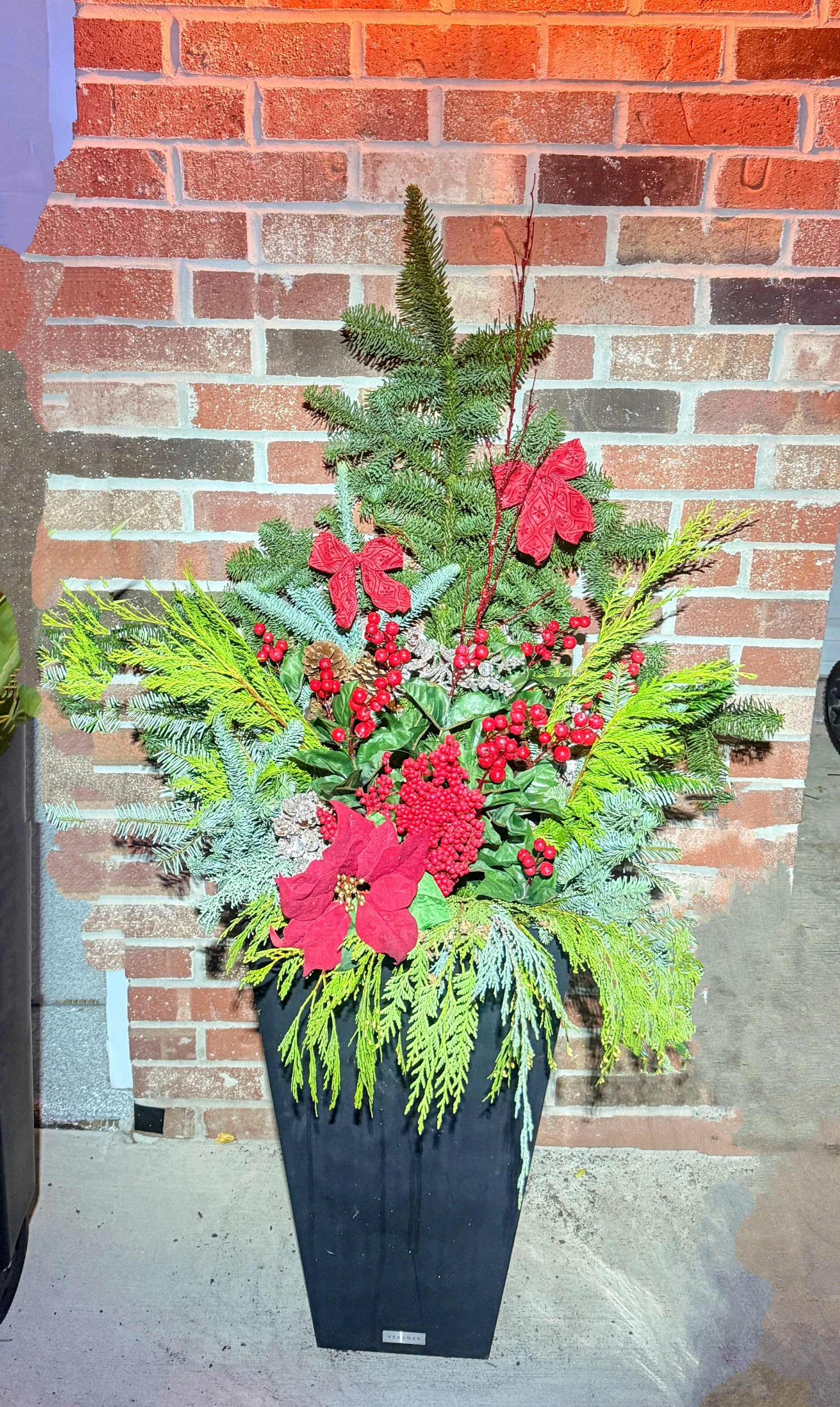 Holiday floral arrangement in a tall black vase with evergreen branches, red poinsettias, red berries, and decorative red bows, placed against a red brick wall.