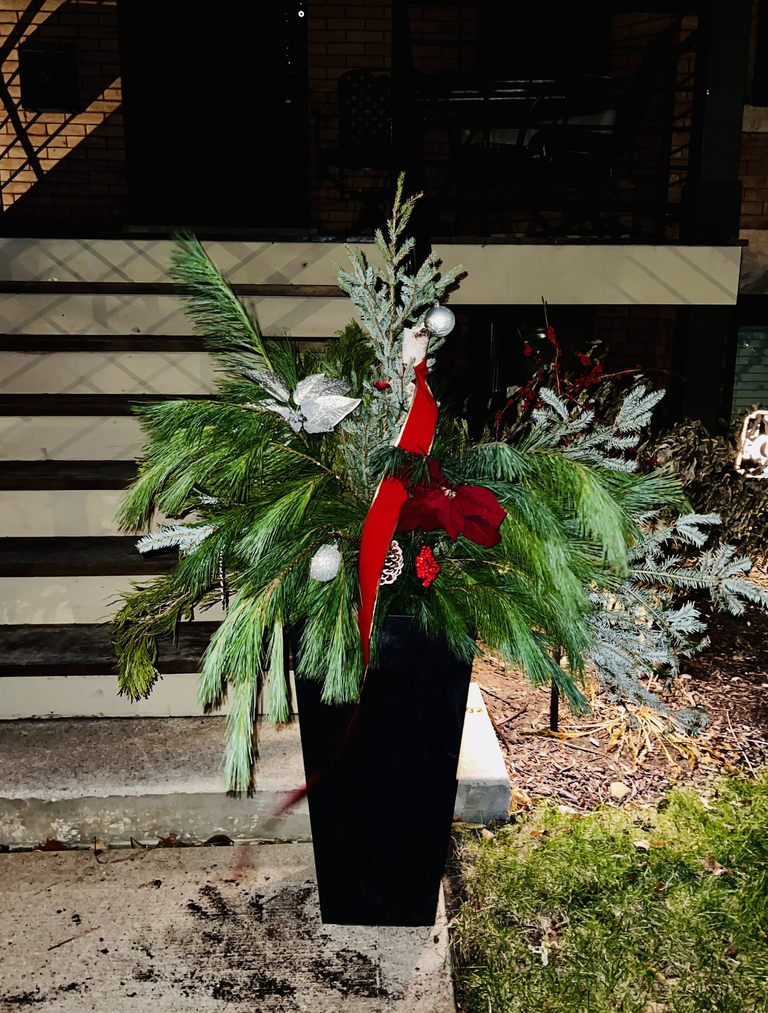 Decorative Christmas floral arrangement in a black vase with green pine and silver frosted leaves, red ribbons, and ornaments, placed outside near a staircase.