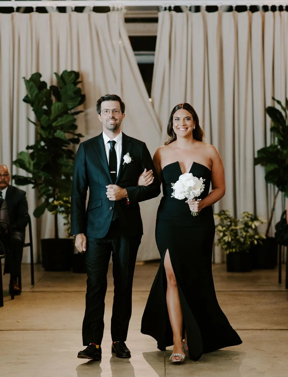 A man and woman in formal attire walking arm in arm at a wedding or formal event. The man is wearing a black suit with a white shirt and black tie, and the woman is wearing a strapless black dress with a thigh-high slit, holding a bouquet of white fl