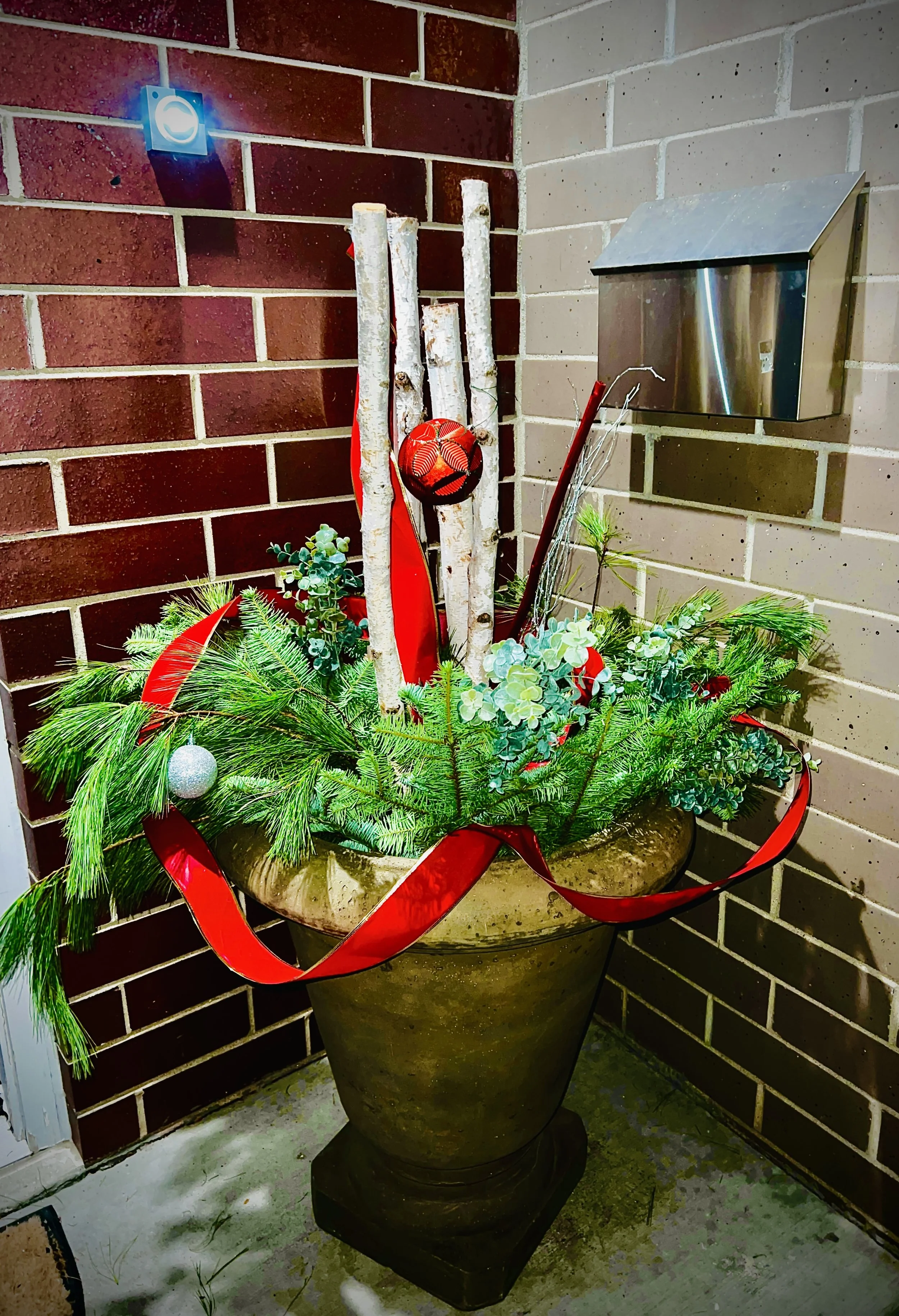 Decorative outdoor pot with holiday greenery, red ribbon, and ornaments, positioned against a brick wall near a mailbox and light.