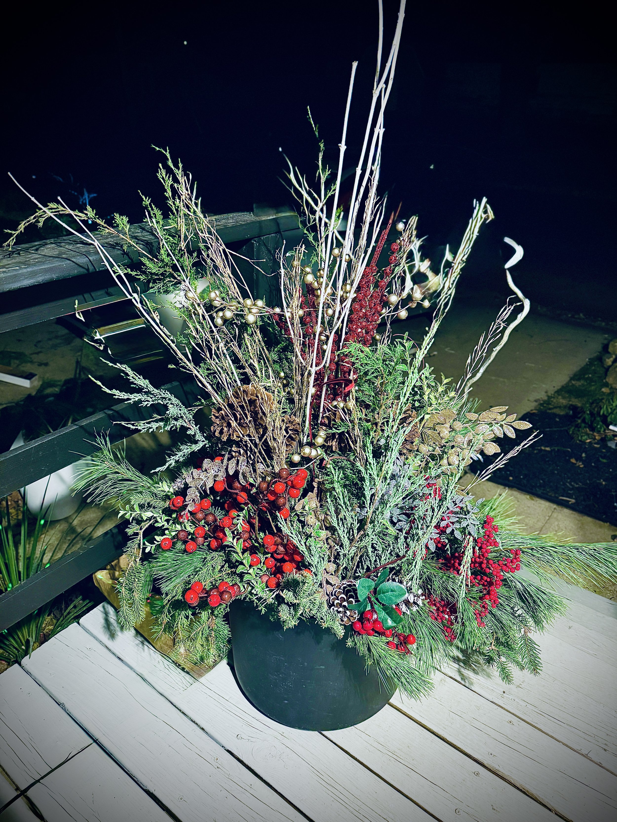 A holiday floral arrangement in a black pot on a white wooden table, featuring pine branches, red berries, pinecones, and decorative gold and red accents, illuminated at night.
