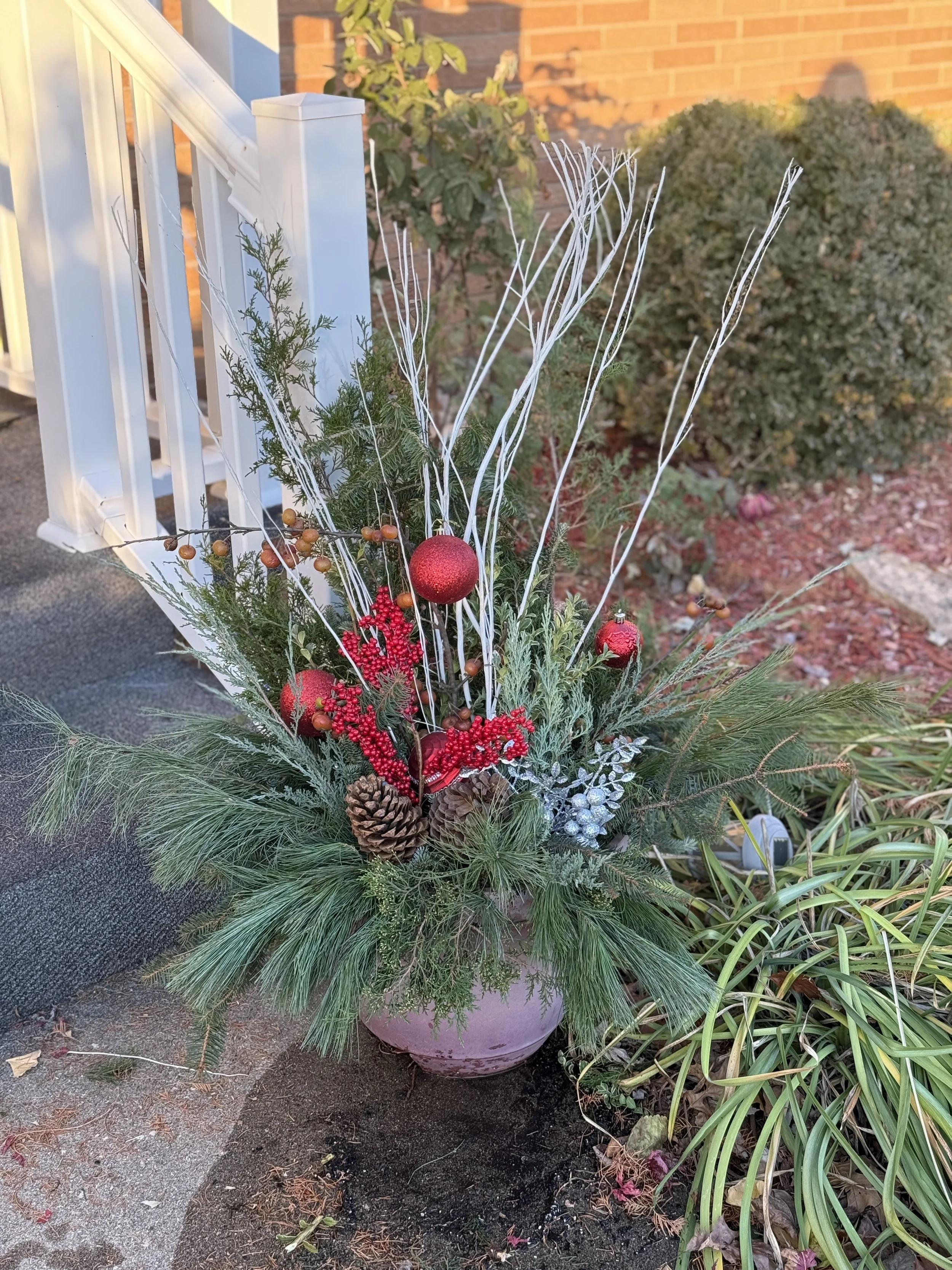 A festive holiday floral arrangement in a purple pot with red ornaments, pinecones, green pine branches, white decorative sticks, red berries, and silver accents placed outdoors next to a brick house and a white staircase.