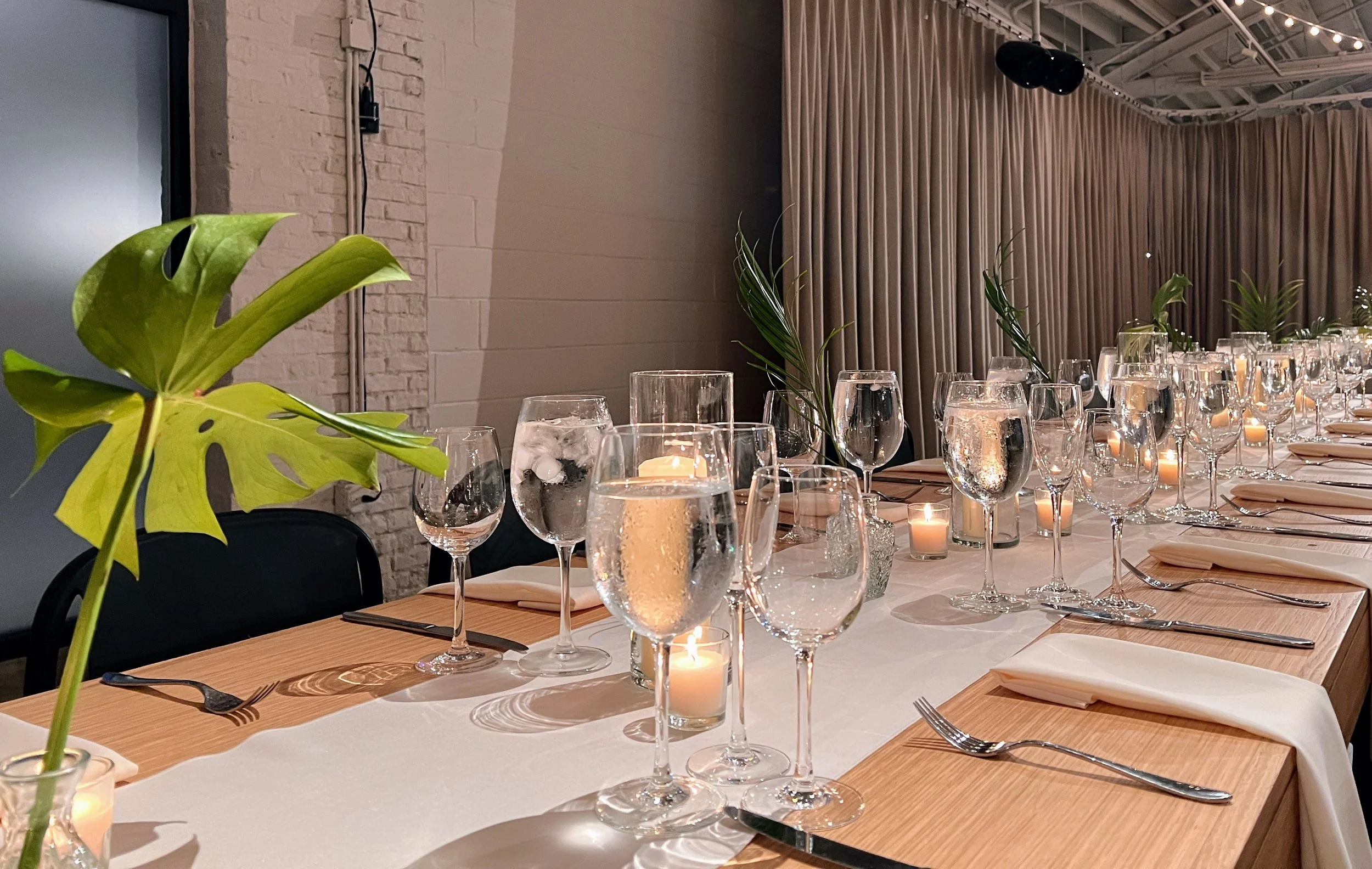 Elegant dining table set with multiple wine glasses, candles, and green foliage, in a modern venue with exposed brick and curtains.