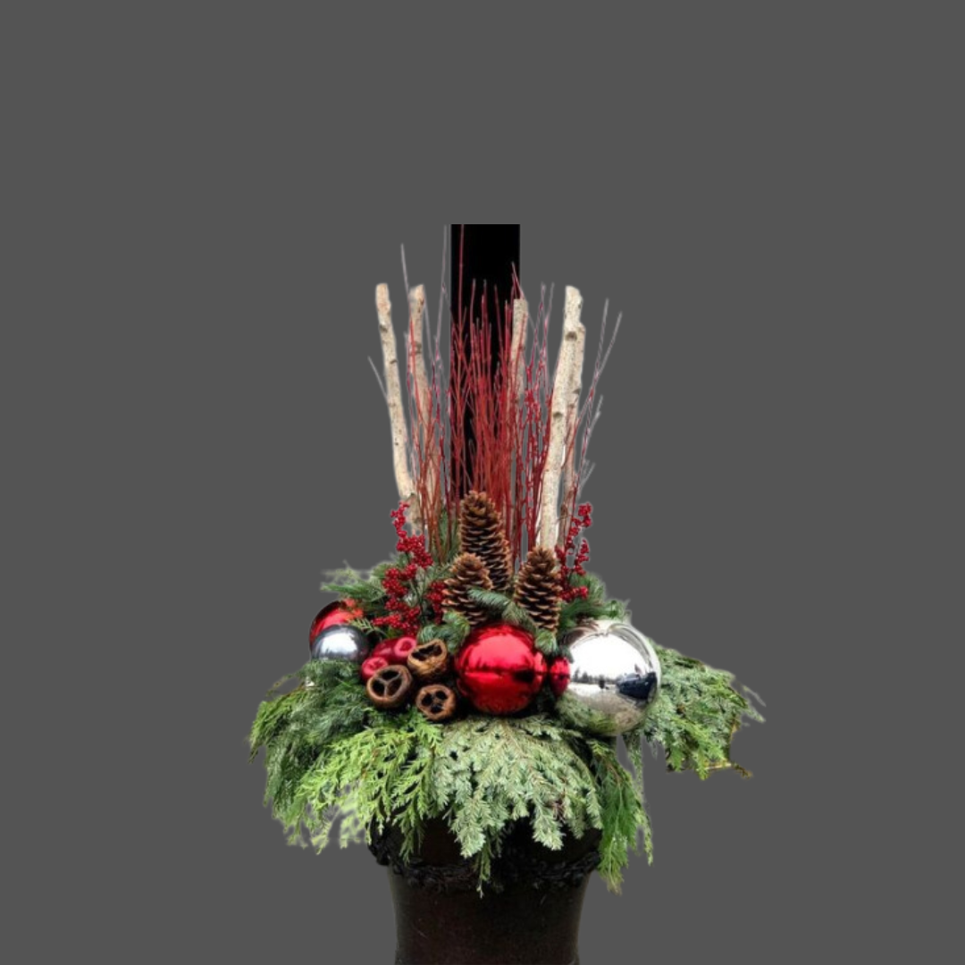 Christmas centerpiece with pine branches, red and silver ornaments, pine cones, cinnamon sticks, red berries, white branches, and decorative stick accents, displayed in a black pot.