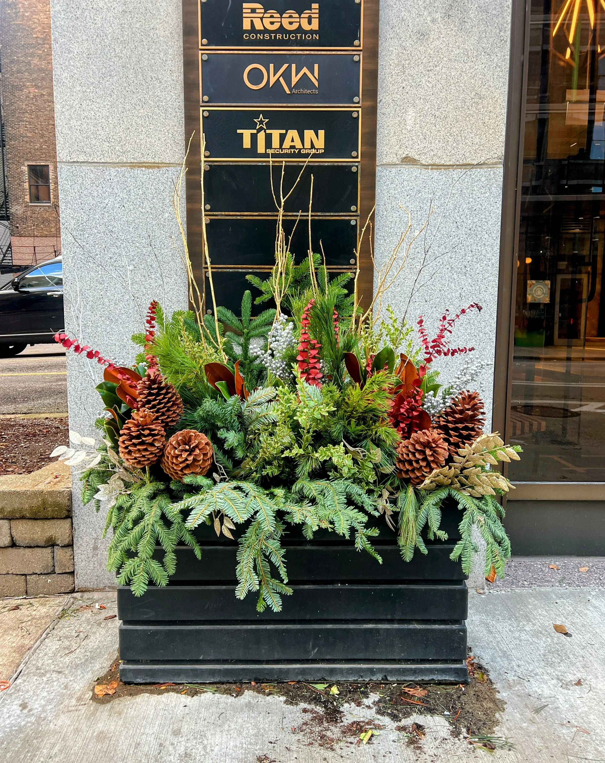 Christmas-themed flower arrangement in a black planter box with pinecones, evergreen branches, red and green foliage, and dried twigs in front of a building with company signs.