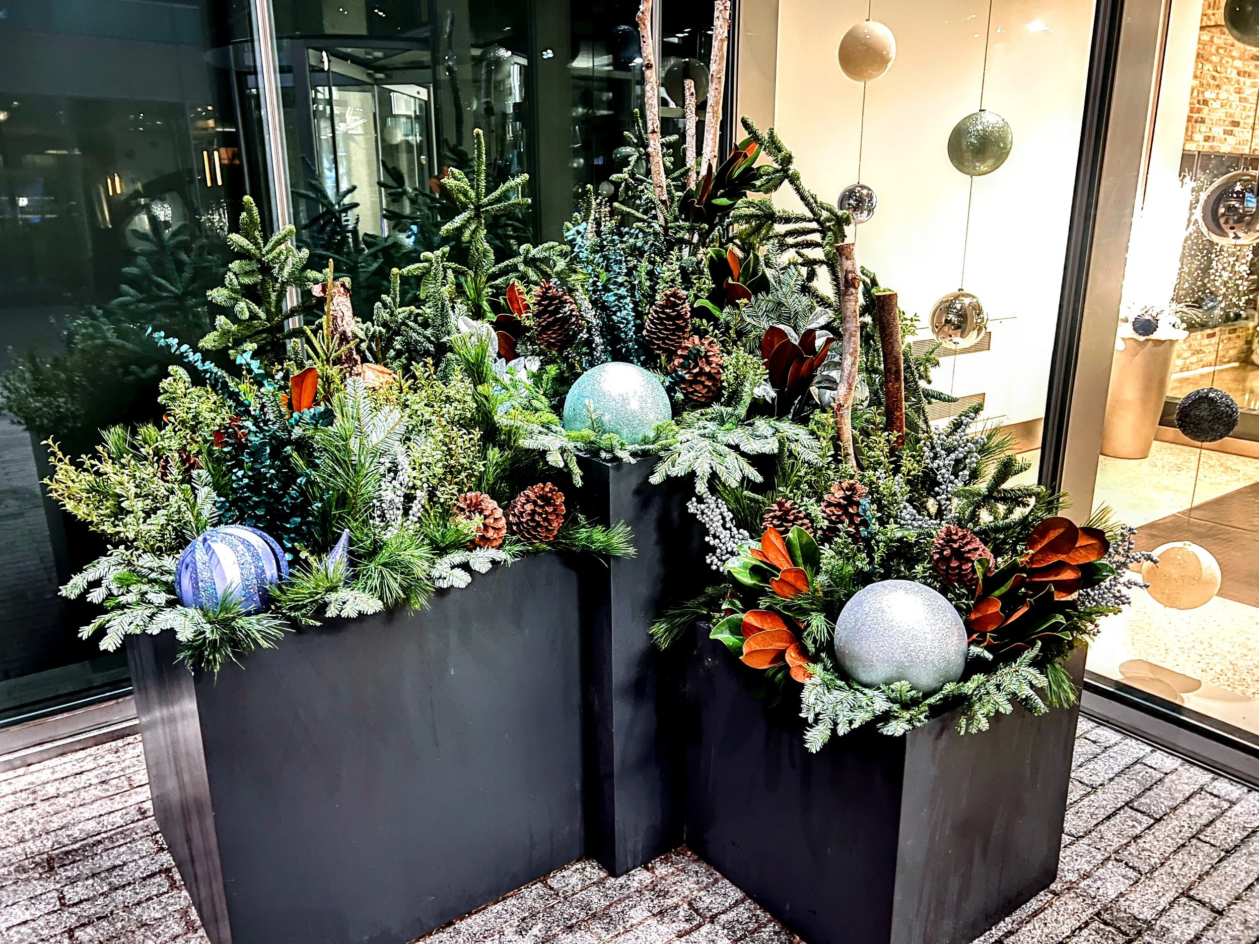 Festive holiday floral arrangement with pine branches, pinecones, and colorful ornaments in black planters near a glass window