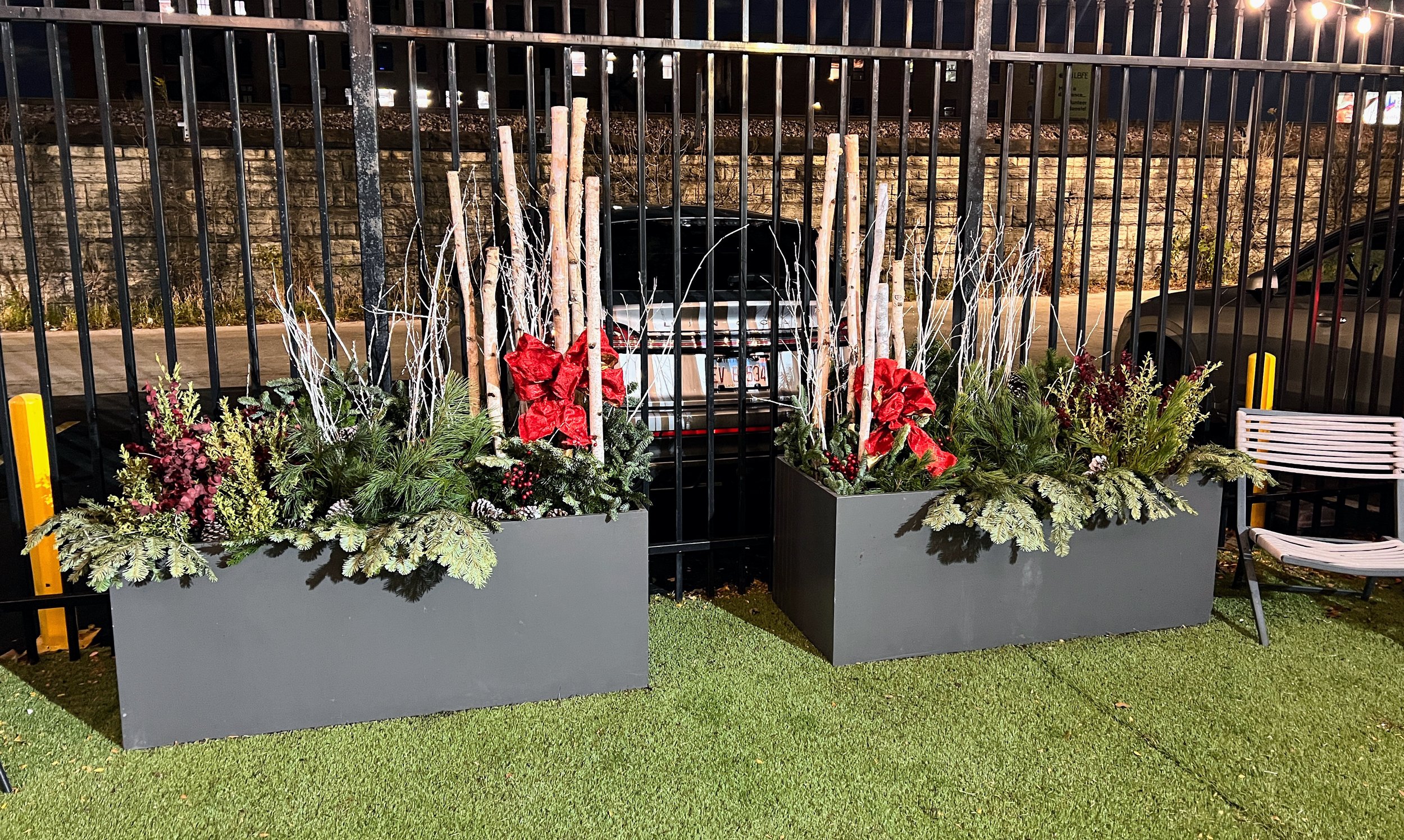 Nighttime outdoor scene with two large decorative planters filled with green plants and red poinsettias, flanked by white twigs, outdoors near a black metal fence, with parked cars and a brick wall background.