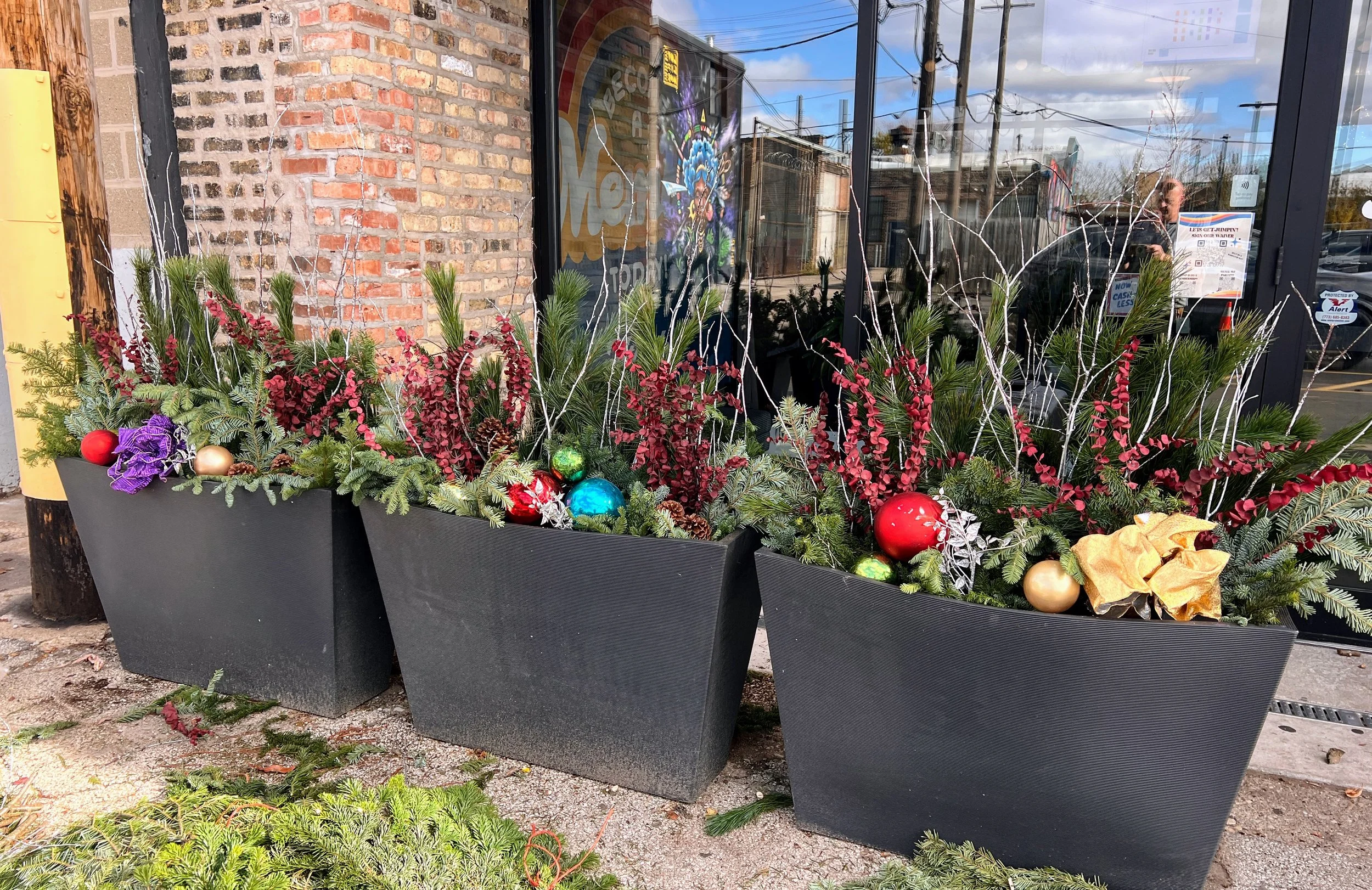 Three large black planters with Christmas decorations, pine branches, red berries, ornaments, and gold and purple elements, placed outside near a brick wall and glass window.