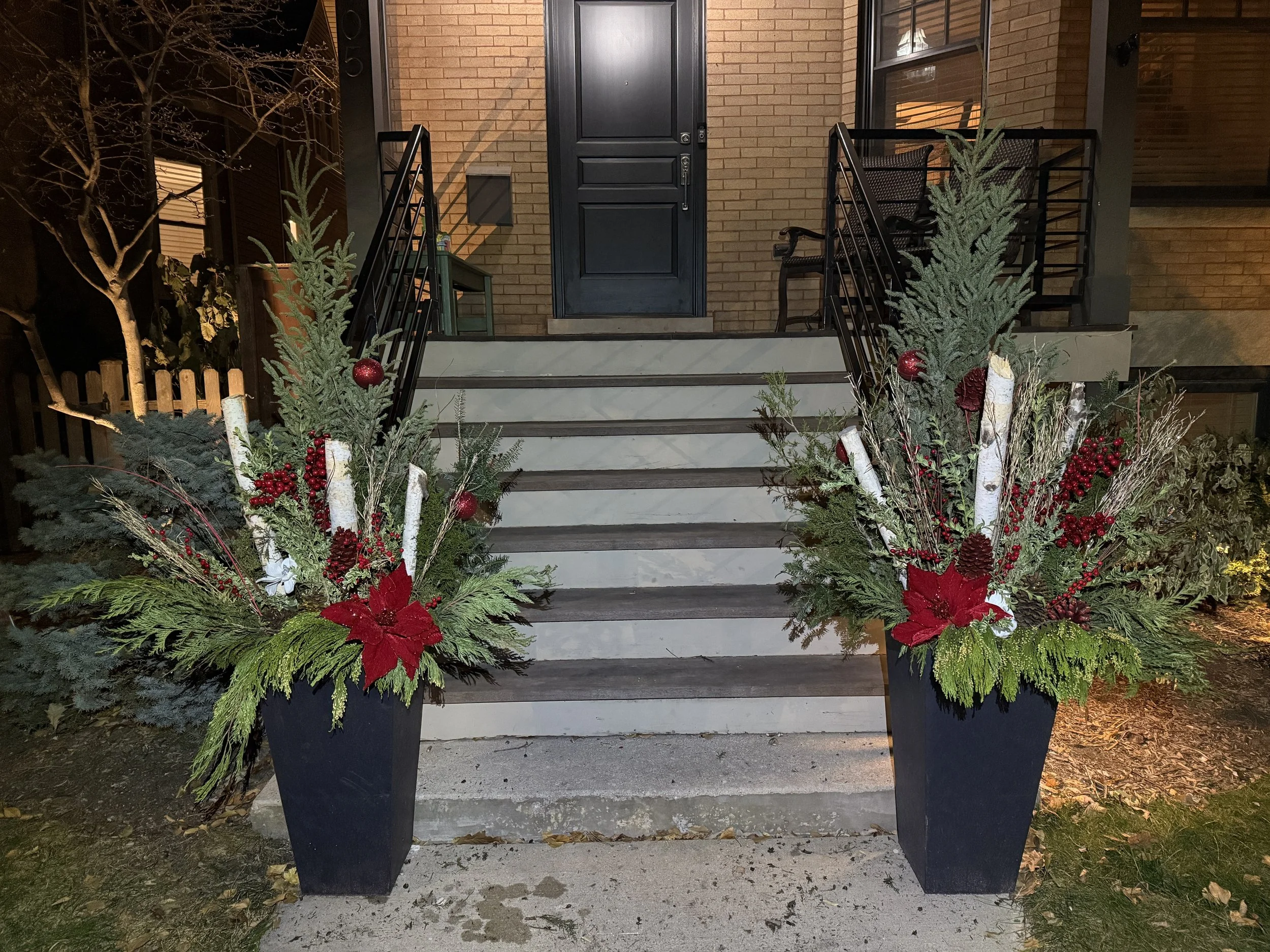 Decorative holiday floral arrangements on black planters flanking front porch stairs leading to a black door, with Christmas ornaments, poinsettias, greenery, and pinecones.