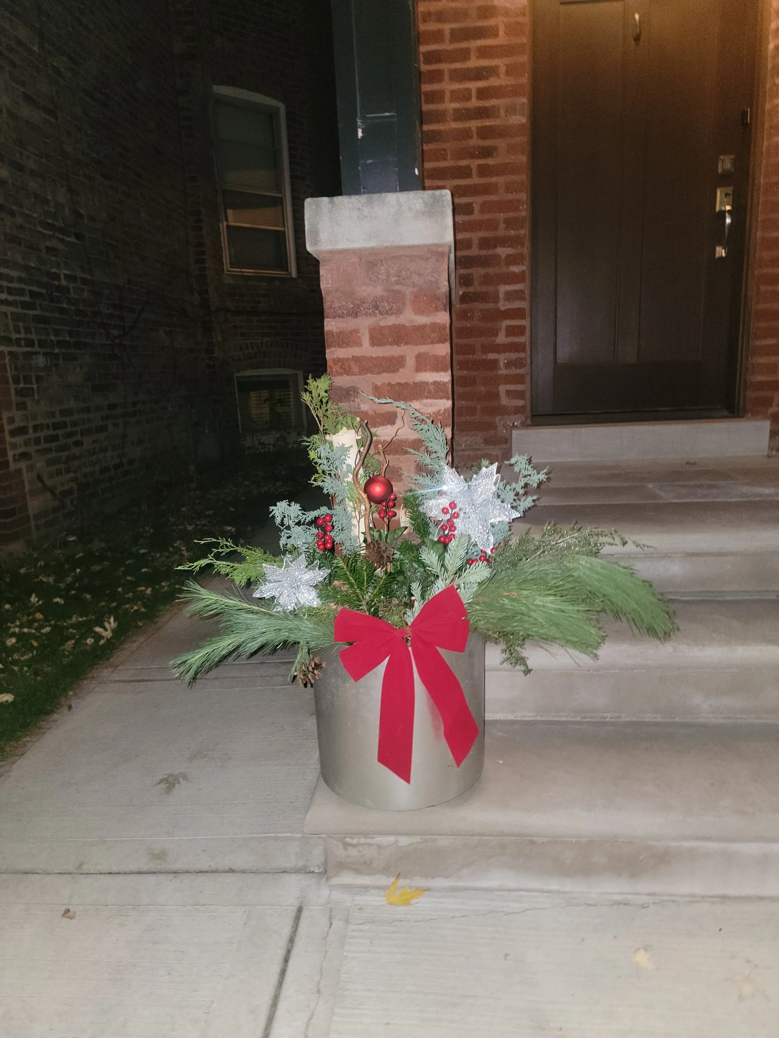 Christmas floral arrangement in a silver pot with pine branches, red berries, silver stars, and a red bow, placed on a porch in front of a brick house.