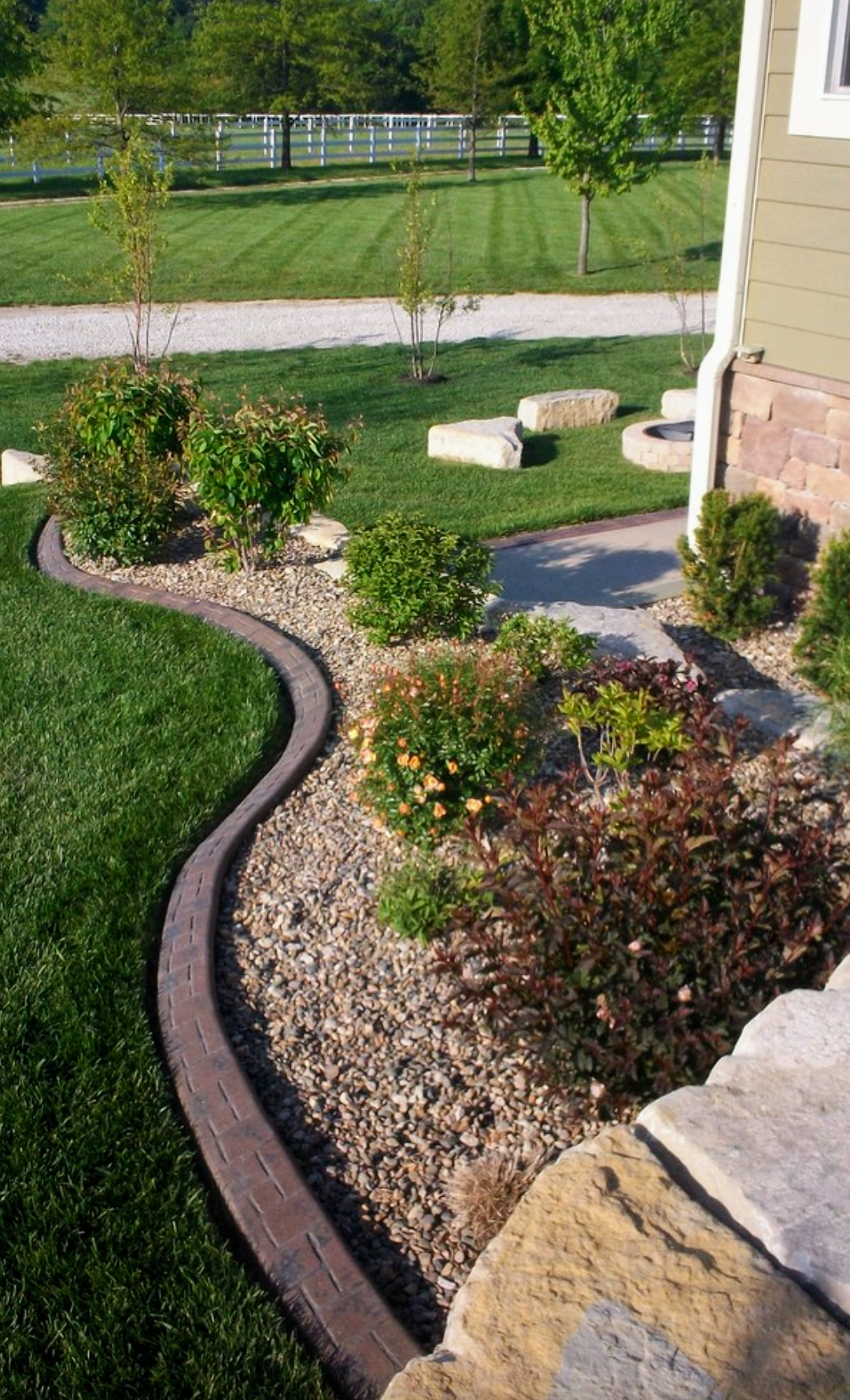 A well-maintained front yard with a curved brick edging separating lush green grass and a garden bed filled with small shrubs and flowering plants, adjacent to a house.