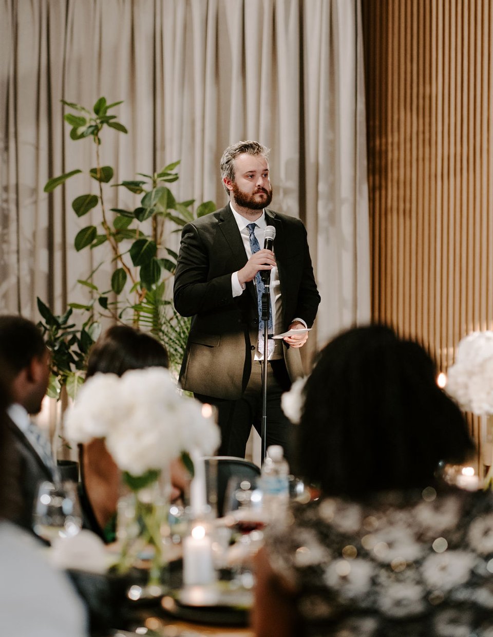 A man in a suit holding a microphone giving a speech at a formal event with floral centerpieces and guests seated at tables.