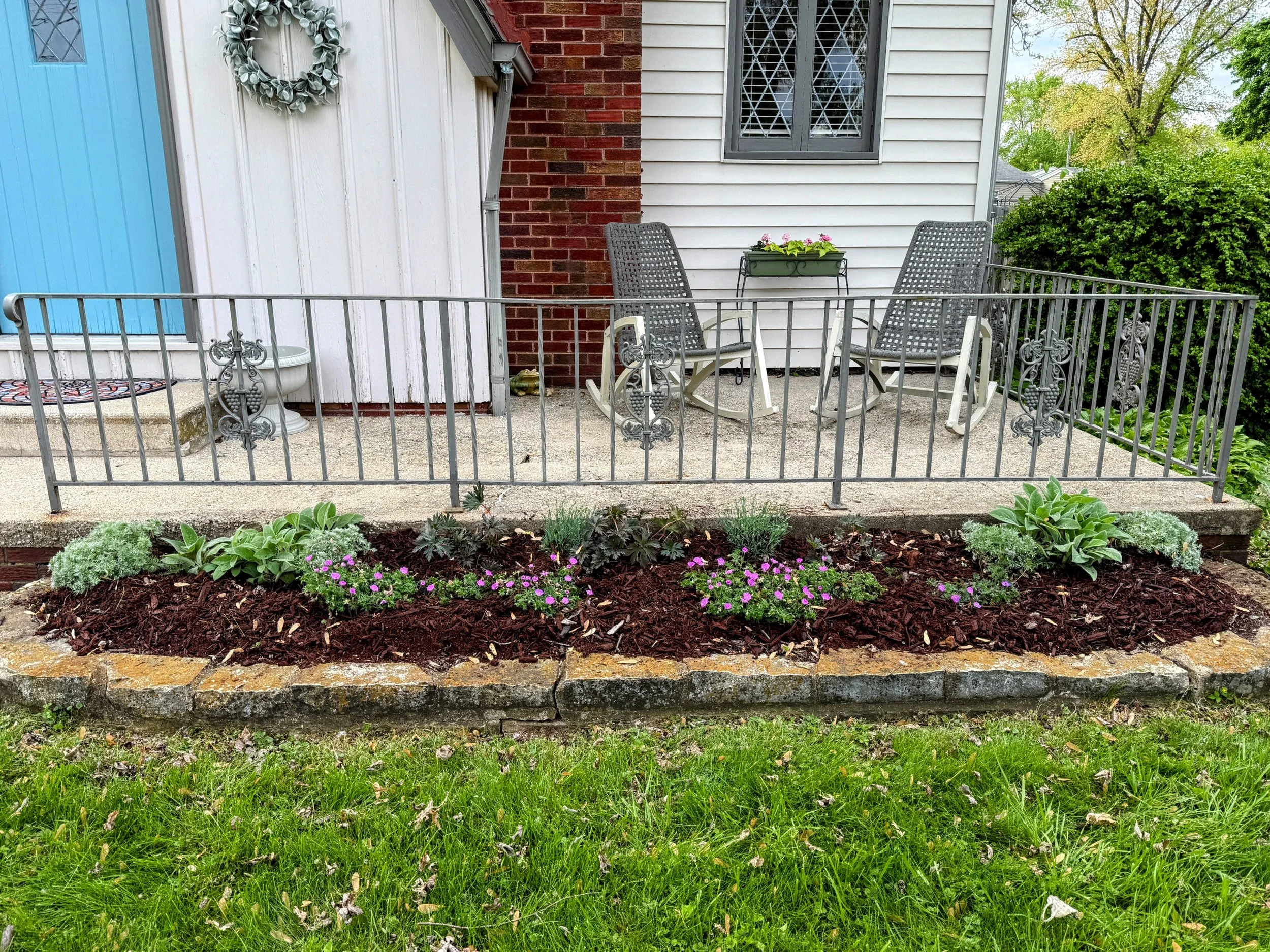 A small backyard patio with a metal railing, two patio chairs, a small glass table, a flower box with pink and purple flowers, and a decorative wreath on the wall. There is a garden bed with green plants and purple flowers in front of the patio, and 