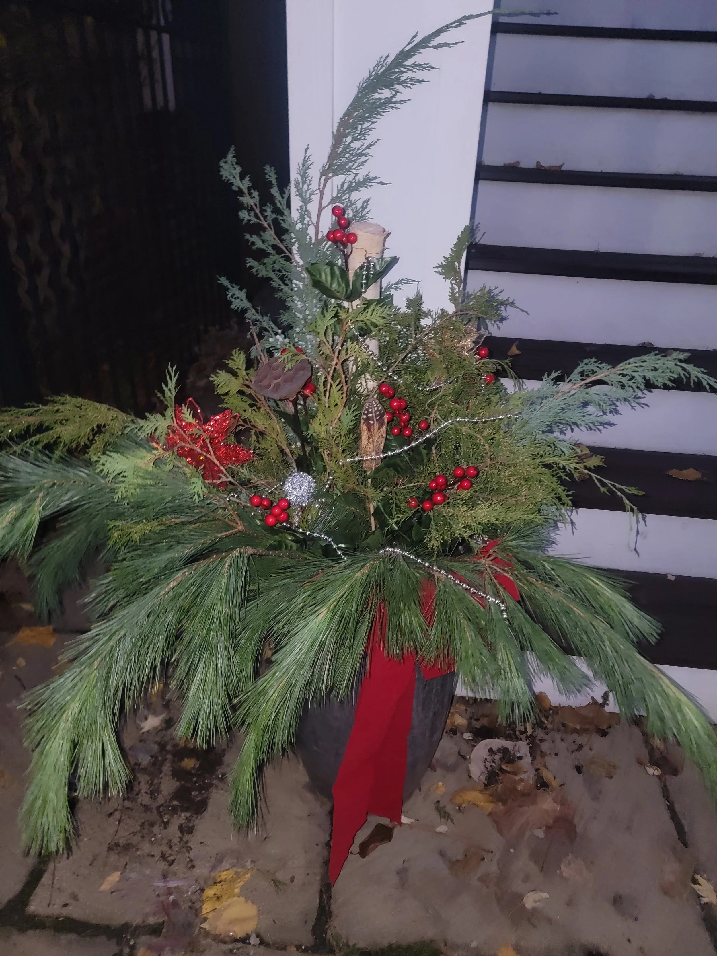 A decorated outdoor Christmas arrangement with pine branches, holly leaves, red berries, and ornaments in a black pot, placed near a white wall and steps.
