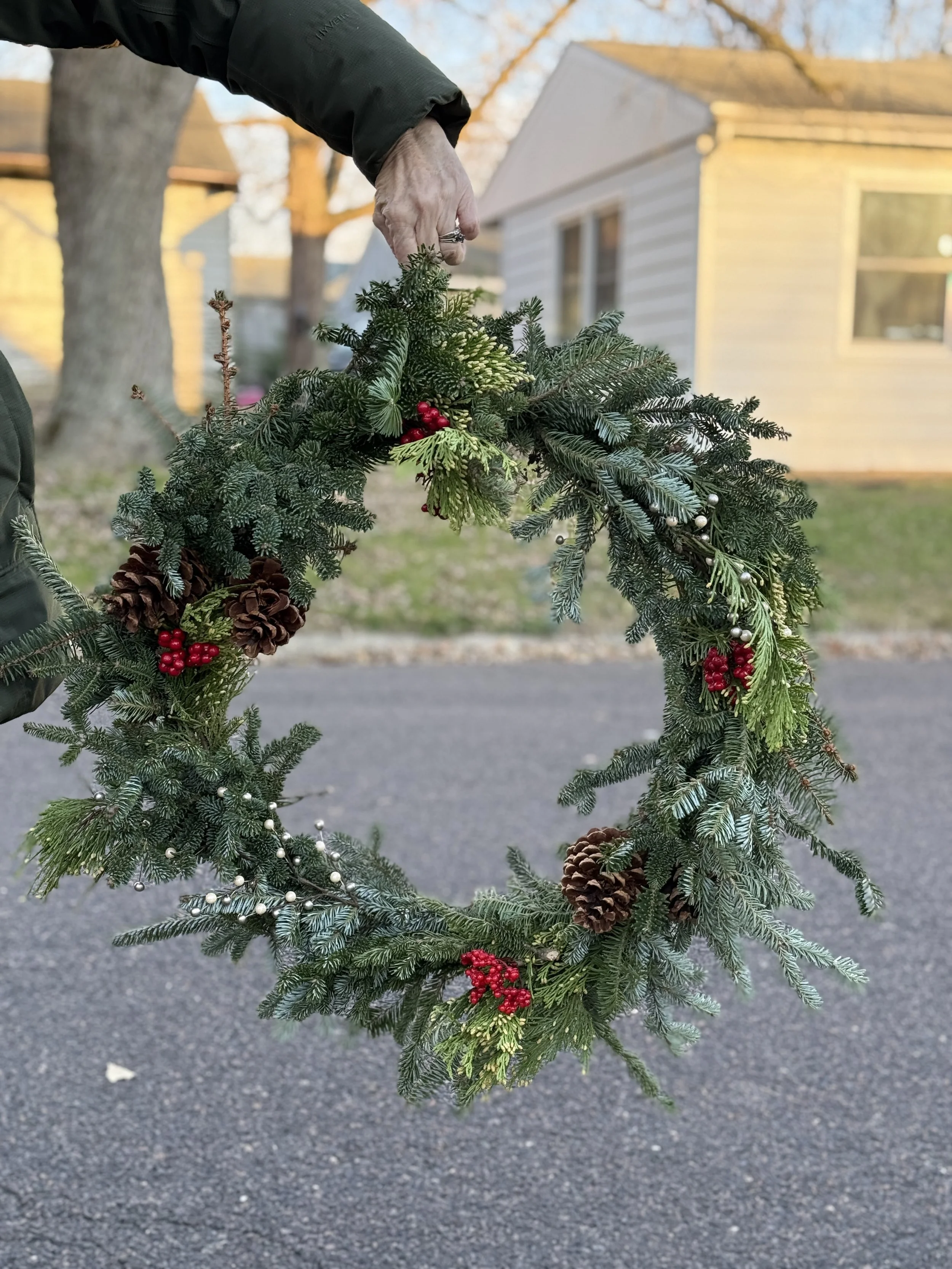 A person holding a decorated Christmas wreath made of pine and fir branches, with pinecones, red berries, white berries, and small ornaments, outdoors with houses and trees in the background.