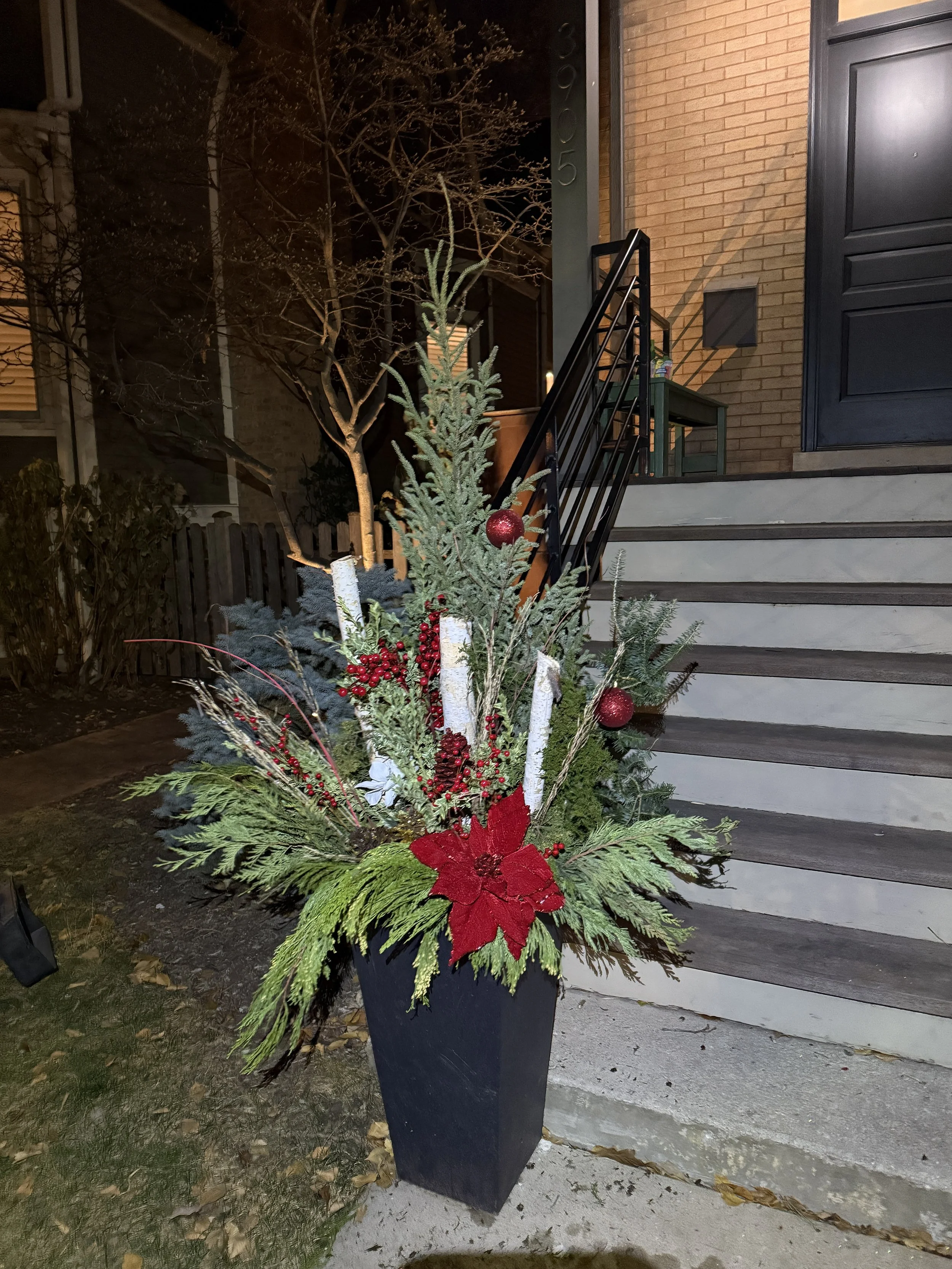 Holiday floral arrangement with evergreen branches, red ornaments, red berries, white candles, a red poinsettia, and pinecones in a tall black planter outside a house at night.