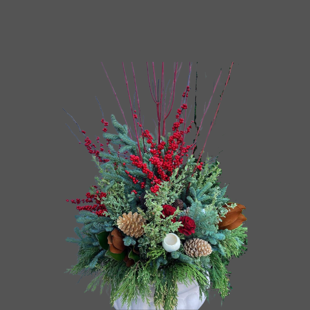 A festive holiday floral arrangement with red berries, red roses, pinecones, evergreen branches, holly leaves, and decorative twigs in a white vase.