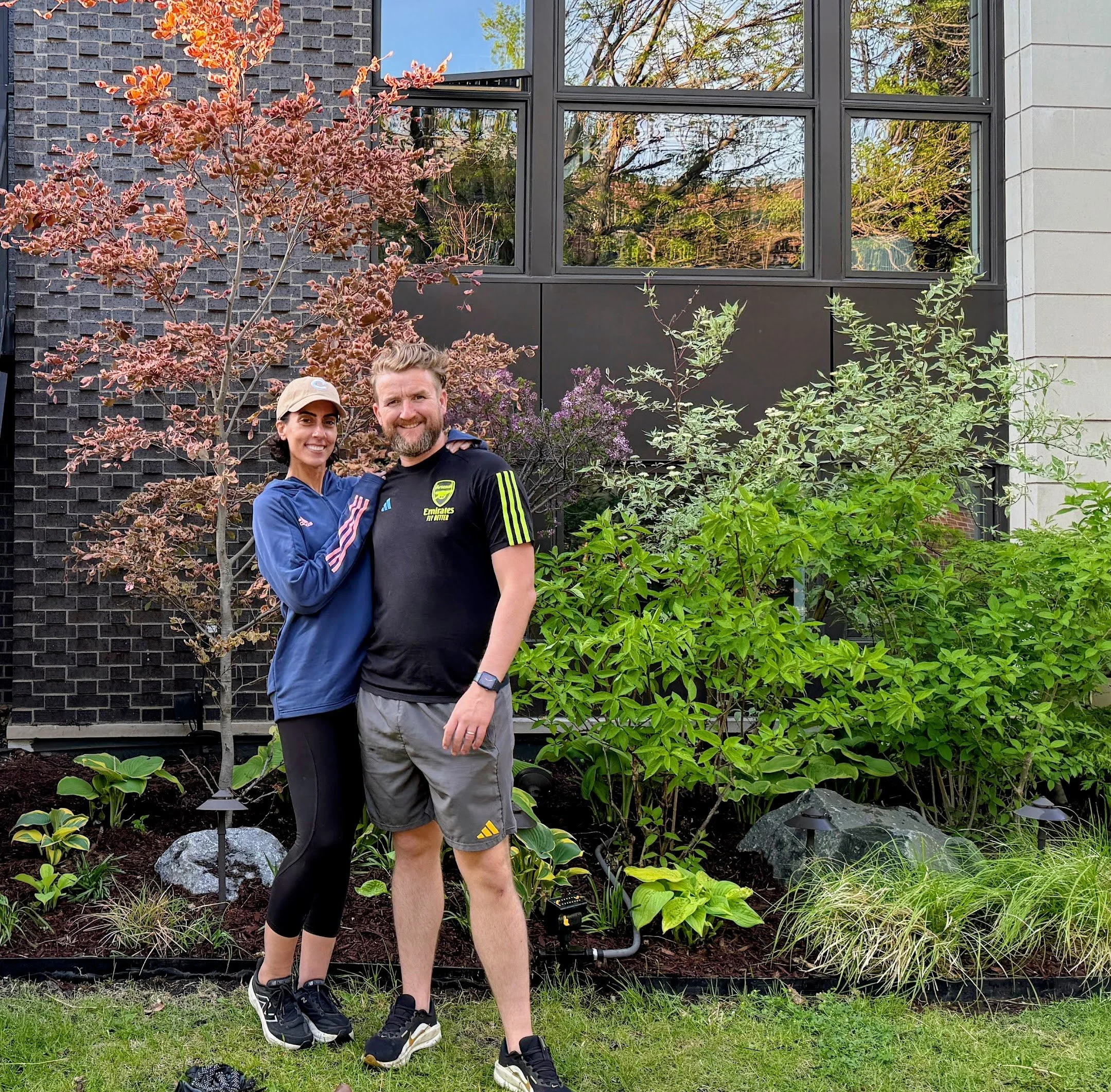 A smiling woman and man standing together in a garden with green plants and trees, in front of a modern home with large windows.